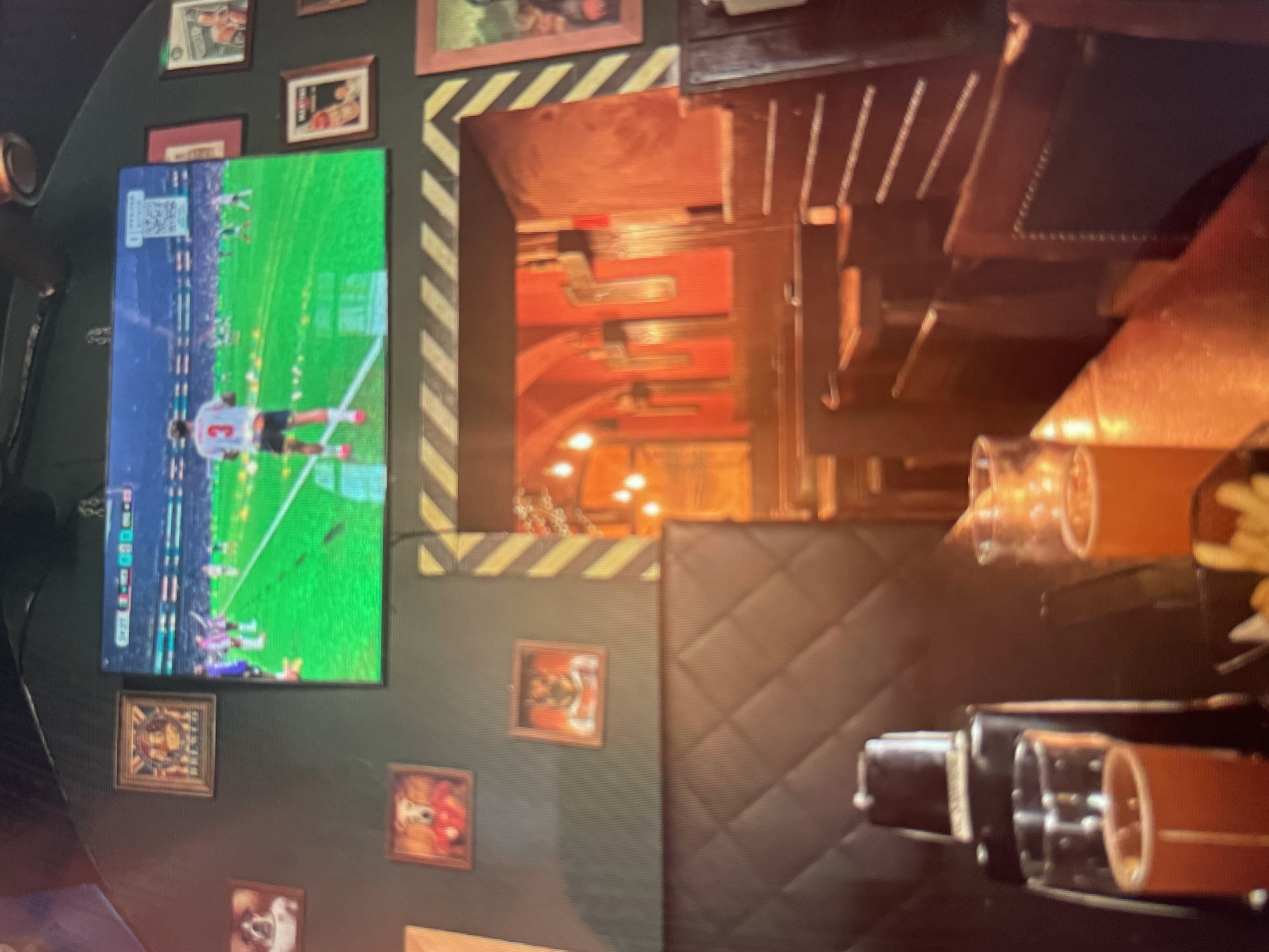 The image depicts the interior of a dimly lit establishment, possibly a bar or restaurant, with a television screen prominently displaying a soccer match. The screen shows players on a green field, with one player in a white jersey with the number 3 on his back. The walls are painted a dark green, adorned with several framed pictures of dogs in clothing, as well as what appear to be vintage advertisements. To the right of the television, a doorway leads into a warmly lit area with red walls and hanging lights. In the foreground, a dark, quilted booth seat is visible. On a table in front of the booth, there is a cocktail shaker and two glasses filled with amber-colored beverages, likely beer or cider. The overall ambiance suggests a casual, perhaps sports-oriented, venue. There is no information in the image to connect it to Shpykolosy, Ukraine.