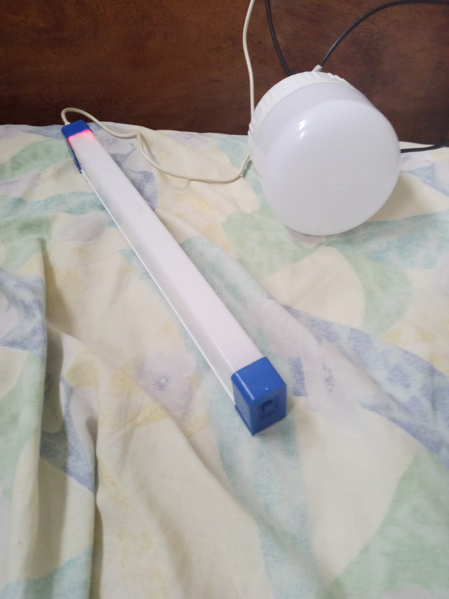 The image shows a white LED tube light with blue end caps and a white round LED light on a patterned fabric surface. A red light is visible at one end of the tube light. The lights are connected to power cords. The background includes a dark wooden surface. The location is Tinaquillo, Venezuela. There are no people visible in the image.