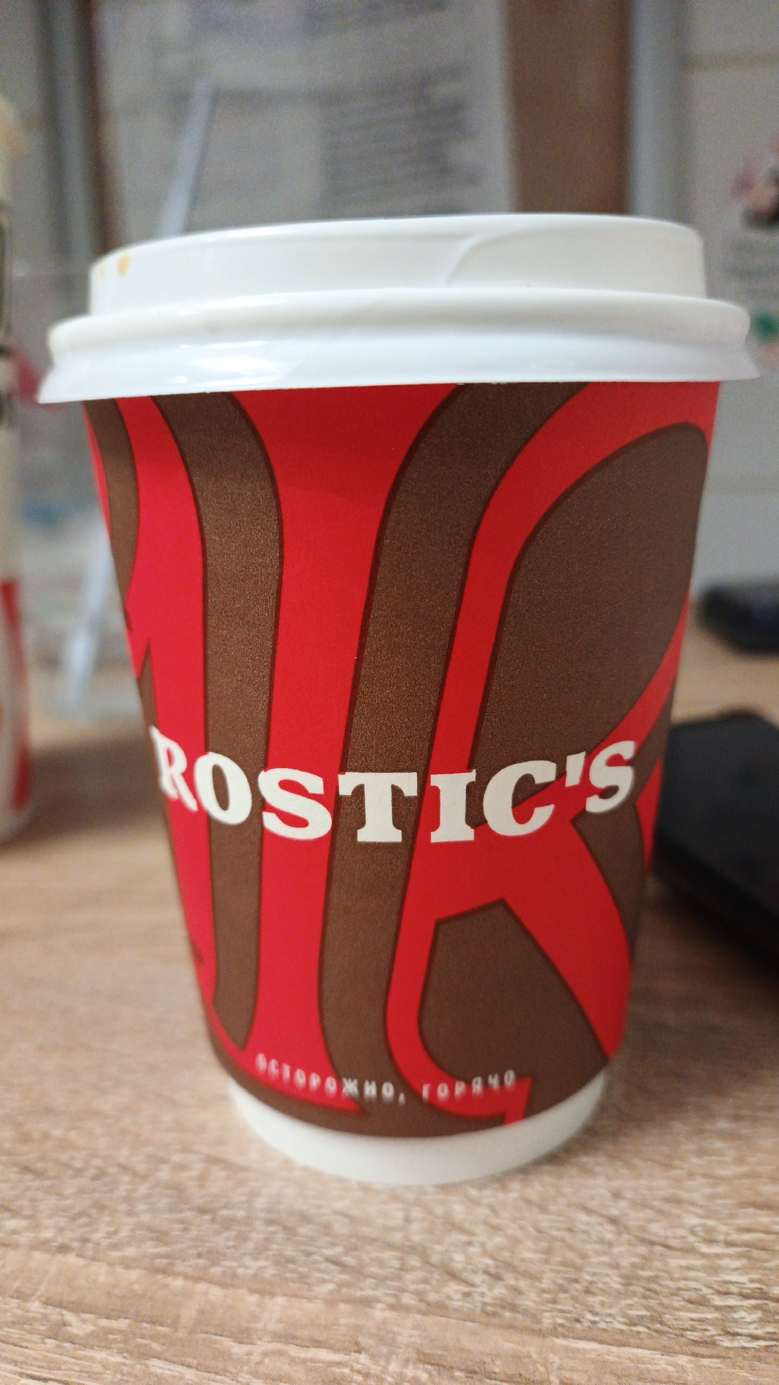 A close-up, eye-level shot shows a red and brown disposable coffee cup with a white lid, placed on a light brown wooden surface. The cup has "ROSTIC'S" printed in white letters on its side, with a red swirl pattern. Below the main text, in smaller white letters, it says "ОСТОРОЖНО, ГОРЯЧО," which translates to "CAUTION, HOT." The background is out of focus, but appears to be an indoor setting with some blurry objects, possibly a desk and a wall. The image does not contain any information about the city of Pavlovsky Posad, Russia.
