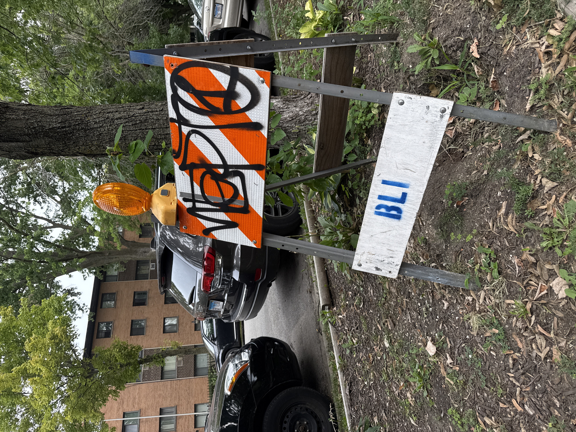 A construction barricade with orange and white reflective stripes stands on a dirt path next to a tree and parked cars. The barricade has graffiti on it and a yellow flashing light on top. A white sign with blue stenciled letters "BLI" is attached to the bottom of the barricade. In the background, a brick apartment building and several vehicles are visible. The environment appears to be an urban residential area.