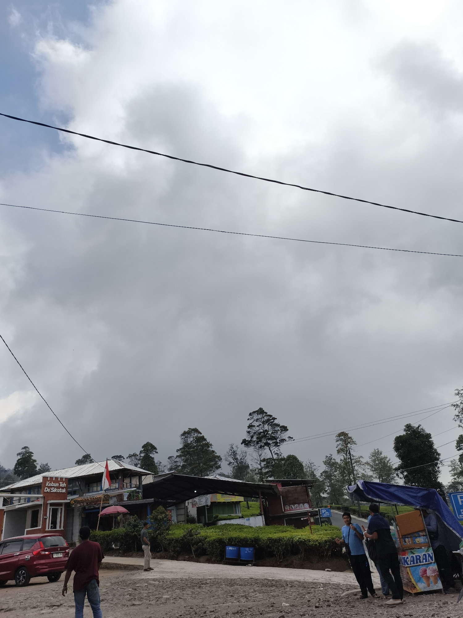 In the city of Sigedang, Indonesia, under a cloudy sky, a scene unfolds by the side of a road. A red car is parked on the left, facing away from the viewer, next to a building with a sign that reads "Kebun Teh De Sikatan". This building appears to be a shop or restaurant, with several people visible inside and outside. Adjacent to it, a food cart displaying images of ice cream and the word "KARAN" is set up, covered by a blue tarp. Three individuals are gathered around the food cart, with one person, a young male wearing a blue shirt, standing and looking towards the viewer, seemingly interacting with the others. Another man in a dark shirt and pants stands next to the cart, while a third man in a light-colored shirt and pants is visible further down the road. Several trees line the background, suggesting a rural or semi-rural setting. The ground is covered in dirt and gravel. Electrical wires are prominently stretched across the sky.