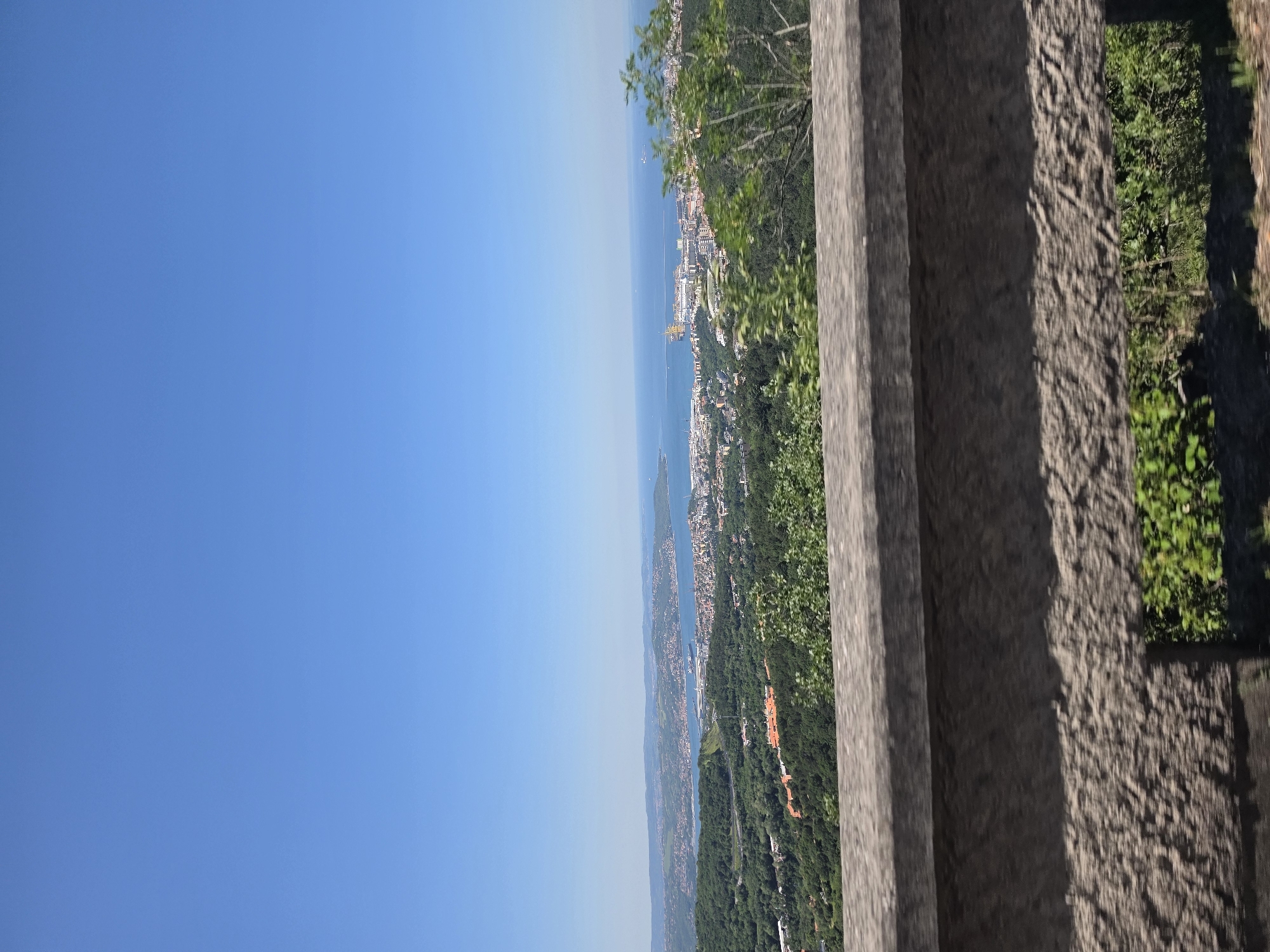 The image shows a view from the Victory Lighthouse (Faro della Vittoria) in Trieste, Italy. The perspective is from behind a stone railing, looking out over the city, the Adriatic Sea, and surrounding forested hills. The sky is clear and blue. The city is densely built up along the coastline. The photo appears to have been taken during the daytime.