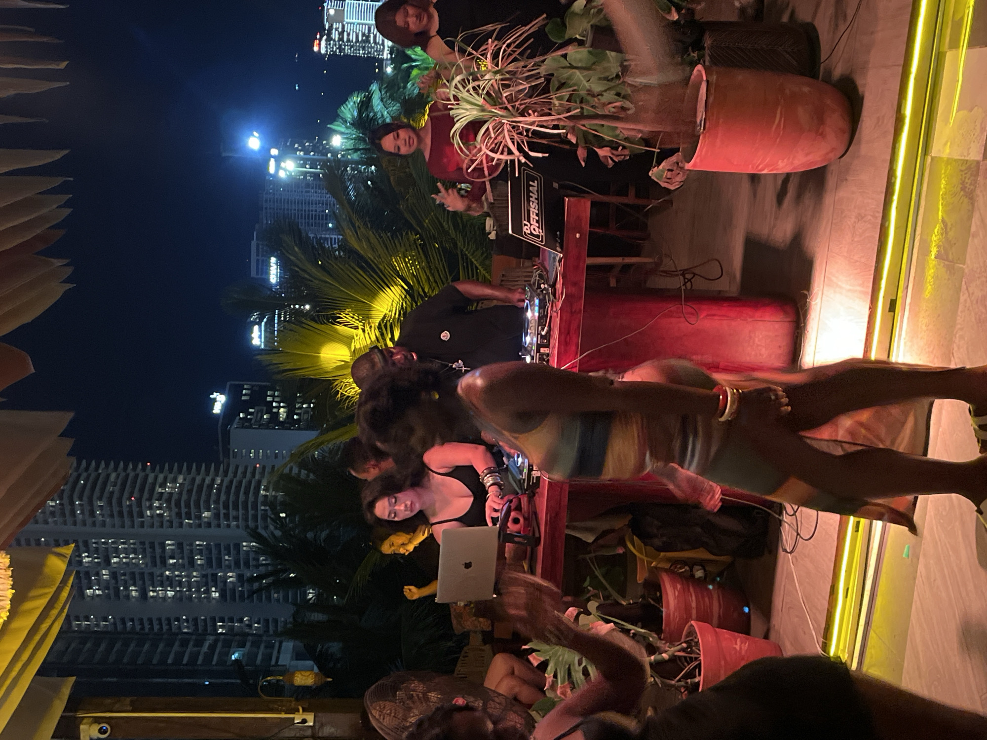 The image depicts a nighttime scene on a rooftop or balcony in Đà Nẵng, Vietnam. In the background, a densely populated cityscape with illuminated high-rise buildings is visible against the dark sky. In the foreground, a group of people are gathered around a DJ setup. Two women are actively engaged with the DJ equipment, with one woman in a black tank top appearing to be operating a laptop and the other in a red top in motion. A male DJ in a black t-shirt and hat is also present at the setup. Several other individuals are present, some appearing to be patrons enjoying the music or socializing. Lush green foliage and potted plants are interspersed throughout the scene, adding a tropical element. The overall atmosphere suggests a lively social gathering or party. The lighting is a mix of ambient city lights, spotlights on the DJ area, and possibly some decorative lighting.