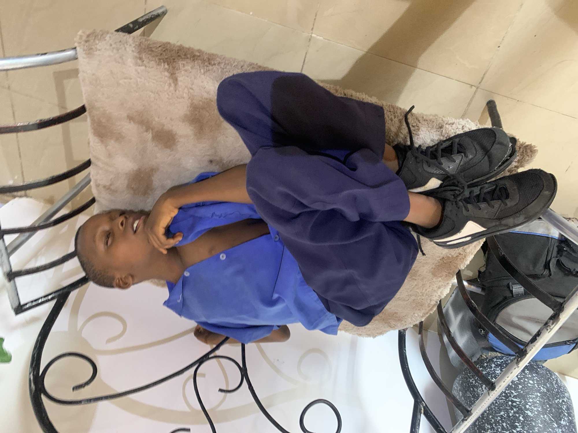 A young person lies on a chair with a light brown cushion. The person wears a blue button-down shirt and dark blue pants, with black sneakers on their feet. The chair has a decorative black wrought-iron frame and rests on a light-colored tiled floor. A backpack is visible beside the chair. The location is possibly in Jos, Nigeria.