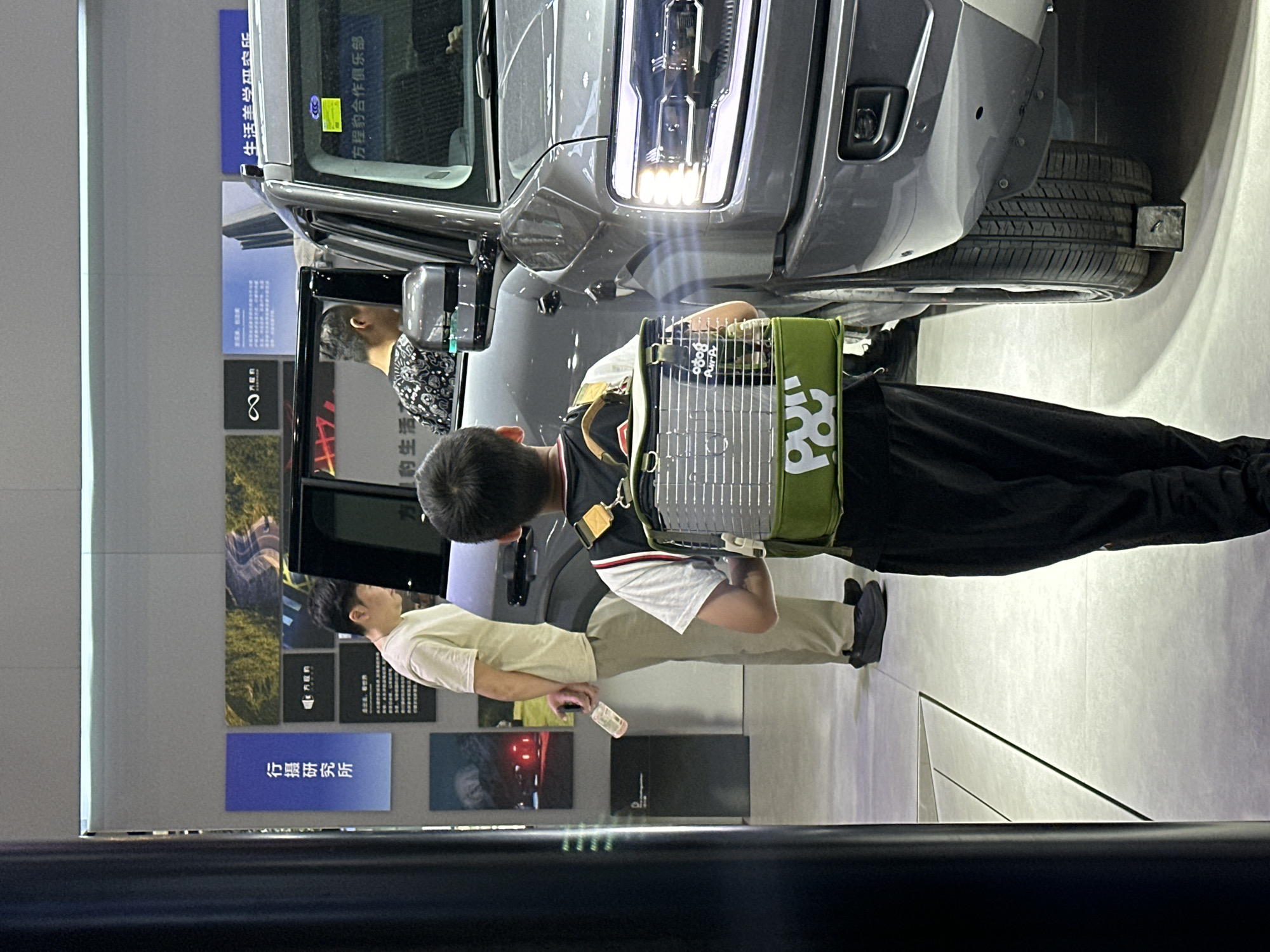 Here is a description of the image.
The image, taken indoors, shows a gray vehicle with its headlights on. The vehicle seems to be the focus of attention for several individuals in the shot.
A young boy, wearing a black and white shirt with black pants, has a green and white backpack-like object on his back. He is standing near the vehicle, possibly examining it. An adult stands next to him.
Behind them, there is a display wall with various photographs and written information, likely related to the vehicle.
In the upper left corner, there are Chinese characters visible, and the text "生活羊学研农乐" and "方程豹合作俱乐部" can be read. The location of "行摄研究所" is also identifiable on one of the panels.
Additional elements in the scene include the presence of other individuals, one of whom is holding a clear bottle. There's a clear view of the vehicle's tires, and the background appears to be a modern exhibition space.