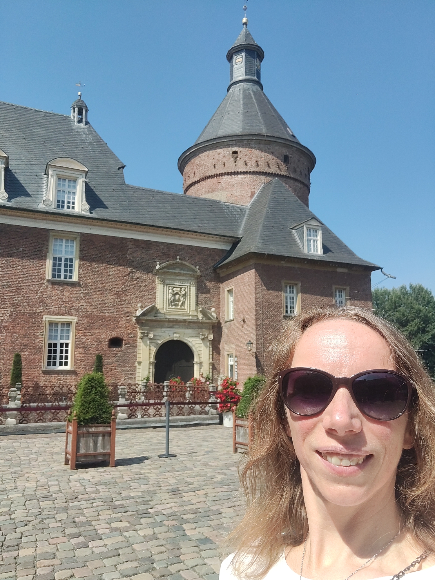 The image shows a woman taking a selfie in front of Isselburg Castle, located in Isselburg, Germany. The castle is a prominent red brick structure with a tall, conical tower. The woman is wearing sunglasses and a white top, and her blonde hair is visible. She is smiling at the camera. The foreground of the image shows a cobblestone courtyard with planters and decorative railings. Trees are visible in the background, and the sky is clear and blue.