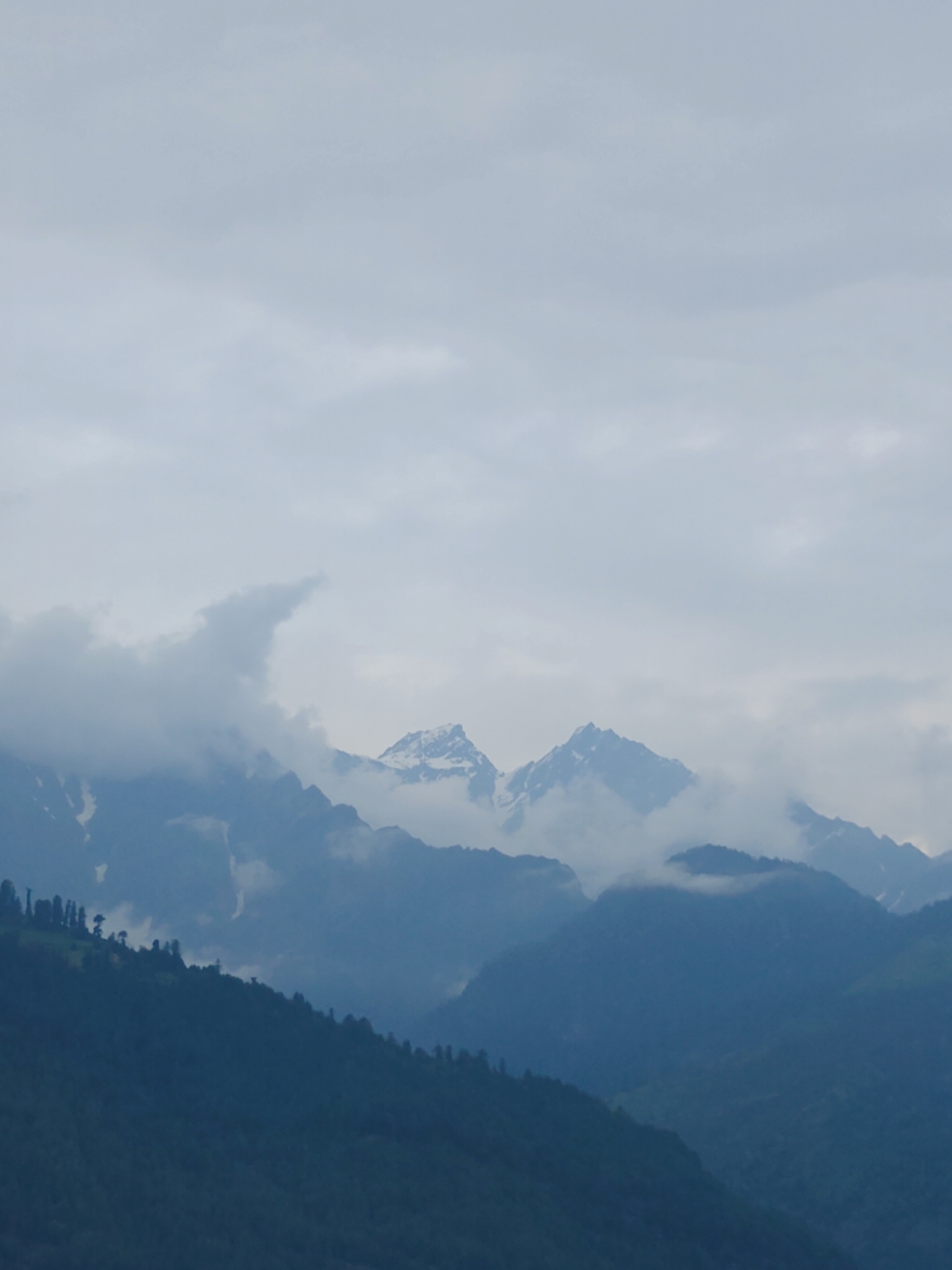 This is a vertical, medium-shot, outdoor photograph of a mountain range under a cloudy sky. The mountains are layered, with darker, forested slopes in the foreground and lighter, snow-capped peaks visible in the distance. Wisps of clouds drift around the higher peaks, obscuring parts of them and creating a sense of depth and mystery. The overall lighting suggests it is daytime, likely overcast or early morning/late afternoon due to the soft light and muted colors. The scene evokes a sense of grandeur and natural beauty, with the imposing mountains and the atmospheric clouds. There is no visible text. The location context provided is Vashisht, India, which aligns with the mountainous terrain and potentially the type of vegetation seen on the slopes. There are no people or specific objects clearly identifiable in the image, making the mountains and the sky the primary subjects. The activity taking place is simply the existence of the landscape, undisturbed and majestic.