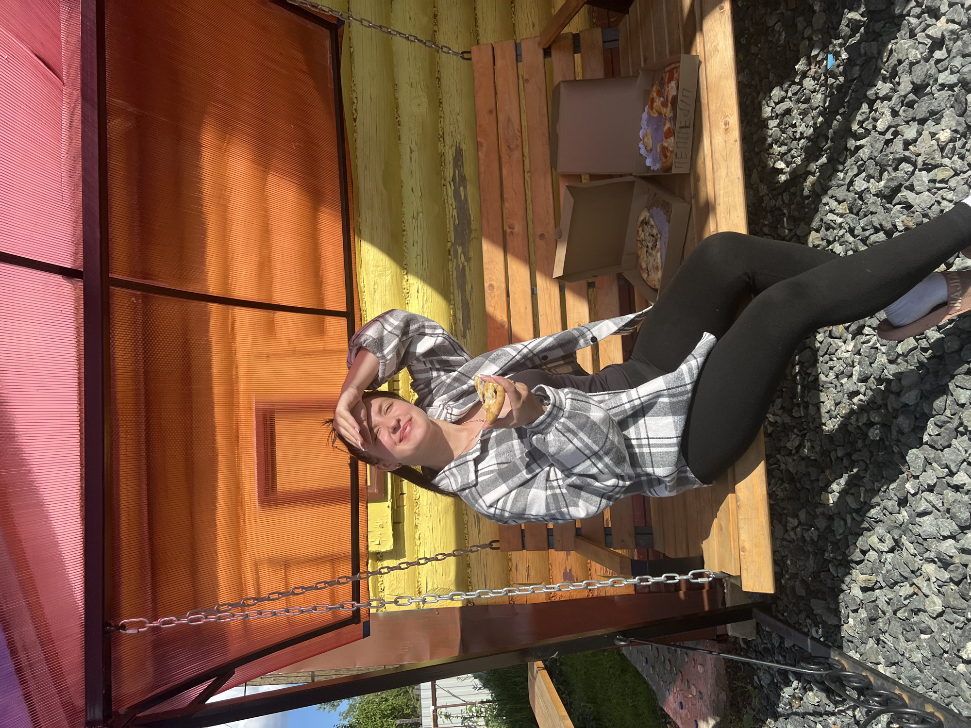 A woman is reclining on a wooden swing set in Noviy, Russia, eating a snack. She wears a plaid shirt and black leggings. Two pizza boxes are situated on the swing set next to her. The swing is suspended from a yellow painted wall and an orange corrugated plastic roof. Gray gravel covers the ground beneath the swing.