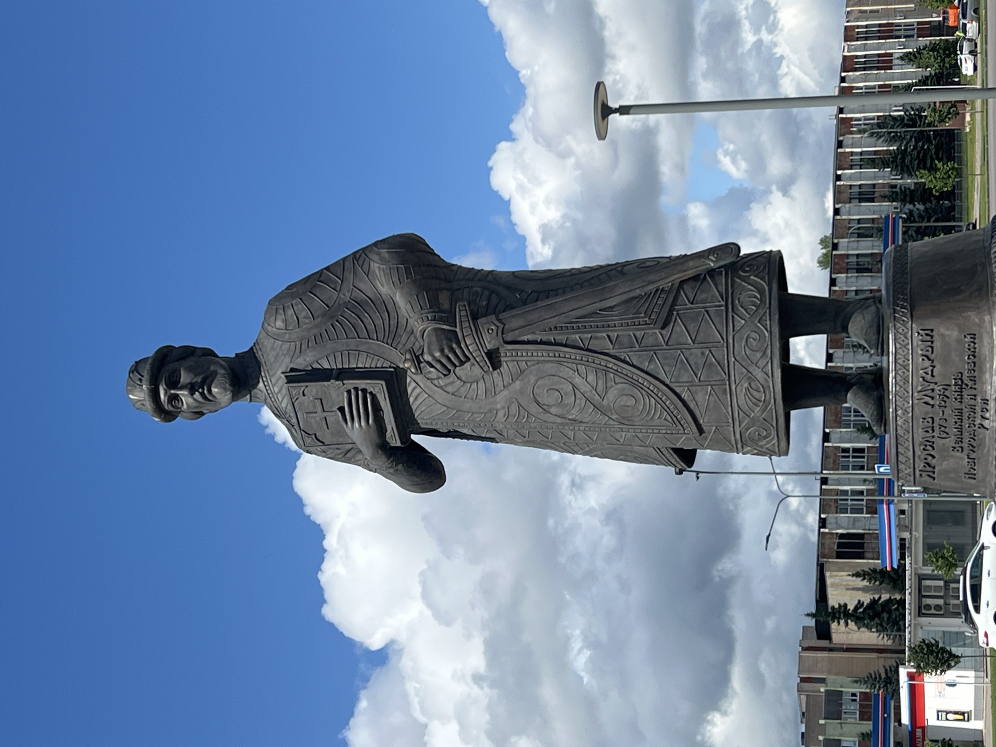 A bronze statue of Yaroslav the Wise stands in the city of Veliky Novgorod, Russia. The statue depicts Yaroslav the Wise holding a book in his left hand and a sword in his right. He is dressed in a long robe with intricate patterns and wears a helmet. The statue is placed on a pedestal with an inscription. In the background, there are several multi-story buildings, trees, and a street with cars. The sky is blue with white clouds. The overall scene appears to be a public monument in an urban setting.