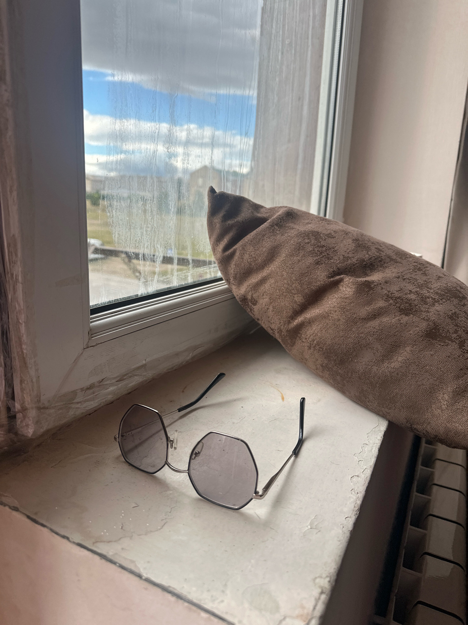 The image depicts a close-up view of a windowsill in what appears to be an apartment in Gyumri, Armenia. On the windowsill, a pair of hexagonal-shaped eyeglasses with thin silver frames and grey lenses are placed. To the right of the eyeglasses, a brown, textured, elongated object, possibly a cushion or bolster, is positioned diagonally, partially obscuring the window. The window itself is dirty, with streaks of water or grime, revealing a cloudy sky and some distant buildings. The windowsill is weathered and shows signs of peeling paint. A portion of a radiator is visible on the right side of the frame. The overall impression is one of a lived-in space, with ordinary objects placed casually.