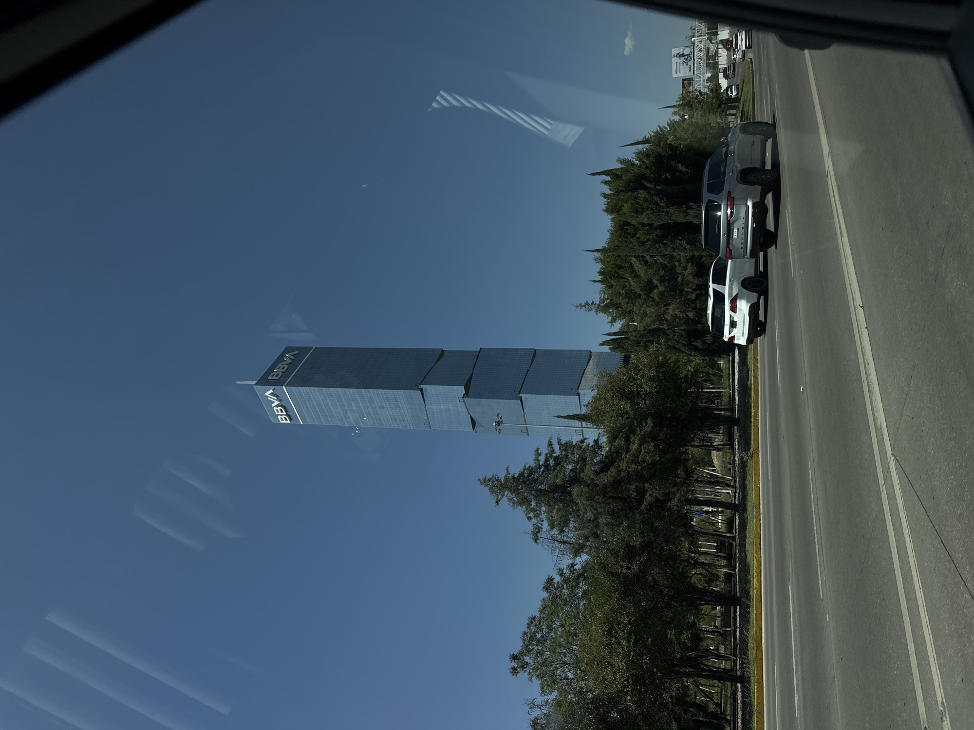 The image shows a view from inside a vehicle, likely a car, looking out at a cityscape. The BBVA skyscraper is prominently featured on the left side of the frame, indicating the location is likely in or near San Bernardino Tlaxcalancingo, Mexico, where the building is located. The sky is clear and blue. On the right side, a highway stretches into the distance with three cars lined up. Trees line the road between the highway and the skyscraper, adding a touch of greenery to the scene. The image is captured through a window, which may distort the view slightly, and there are reflections visible on the glass.