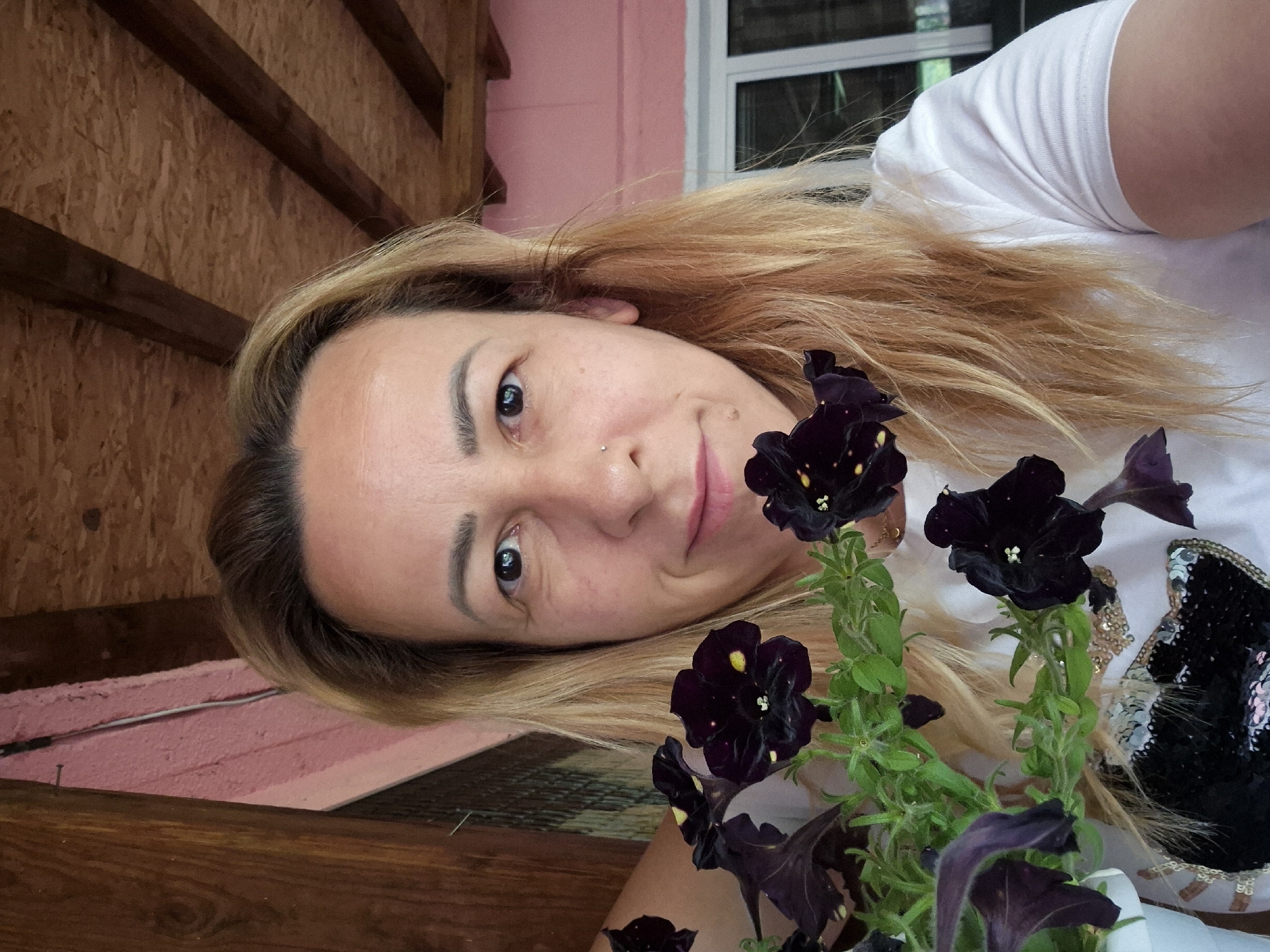 A woman with blonde hair is holding a potted plant with dark purple flowers and yellow centers. She is smiling and looking at the camera. She is wearing a white t-shirt with a sequined design on it. The background shows a pink wall and a wooden ceiling. The scene appears to be outdoors, possibly on a porch or patio.