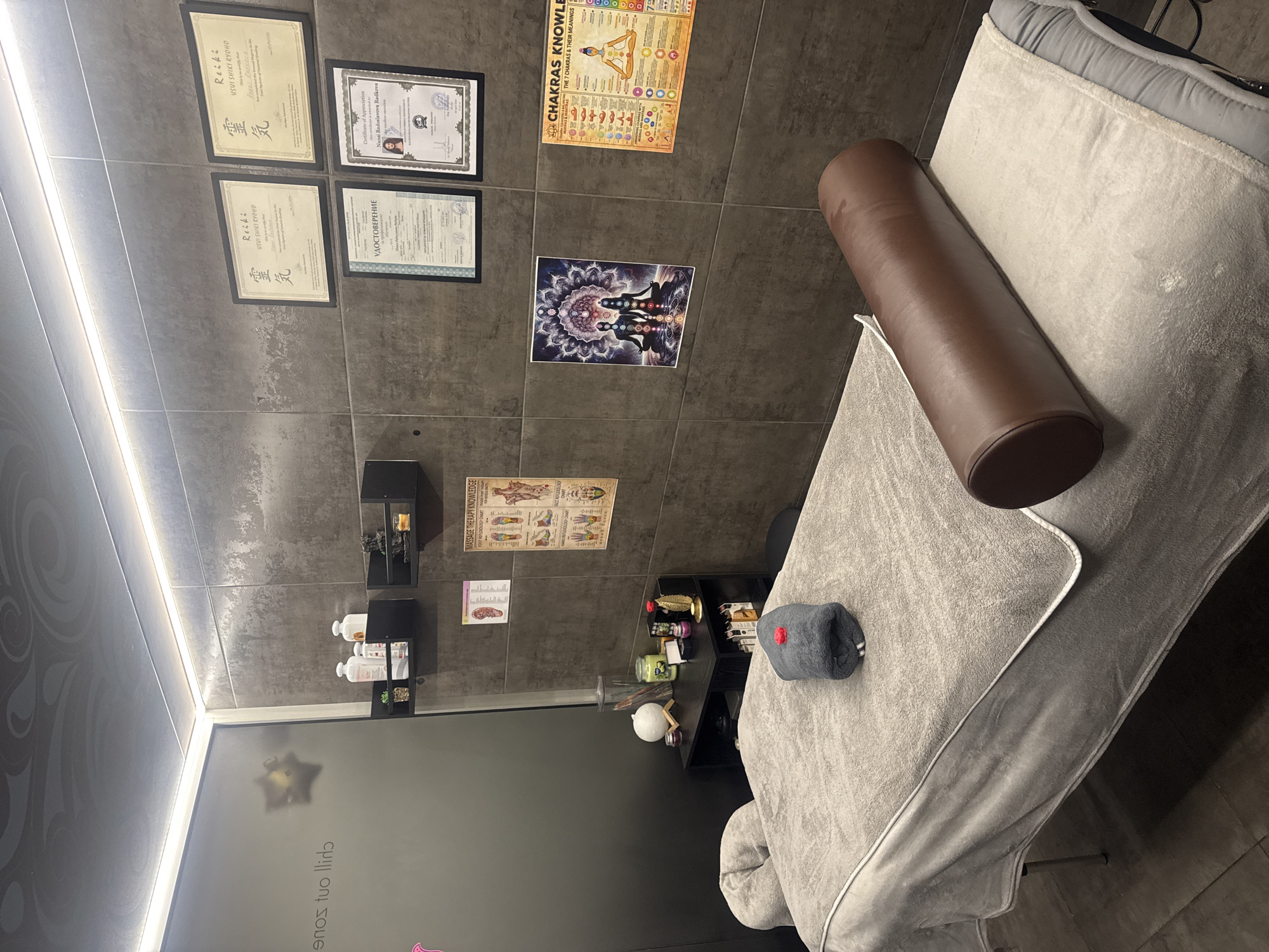 The image shows the interior of a treatment room, likely a massage or therapy center. Certificates and educational posters related to Reiki, chakras, and massage therapy are displayed on the wall. A massage table is covered with a grey sheet, with a brown cylindrical pillow on it. Near the foot of the table is a grey rolled-up towel with a red dot. There's a dark shelving unit with various items like candles, jars, and decorative figures. A glass wall or door partially visible at the bottom left has the phrase "chill out sons" printed on it. Based on the certificate in the frame, it seems like this image was taken in Varna, Bulgaria.