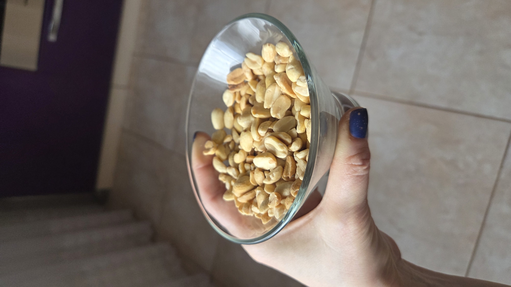 A hand holds a glass filled with peanuts. The hand is fair-skinned with dark blue nail polish. The background shows beige tiled walls and, out of focus, a purple door next to a set of stairs. The location is Chaikovsky, Russia.