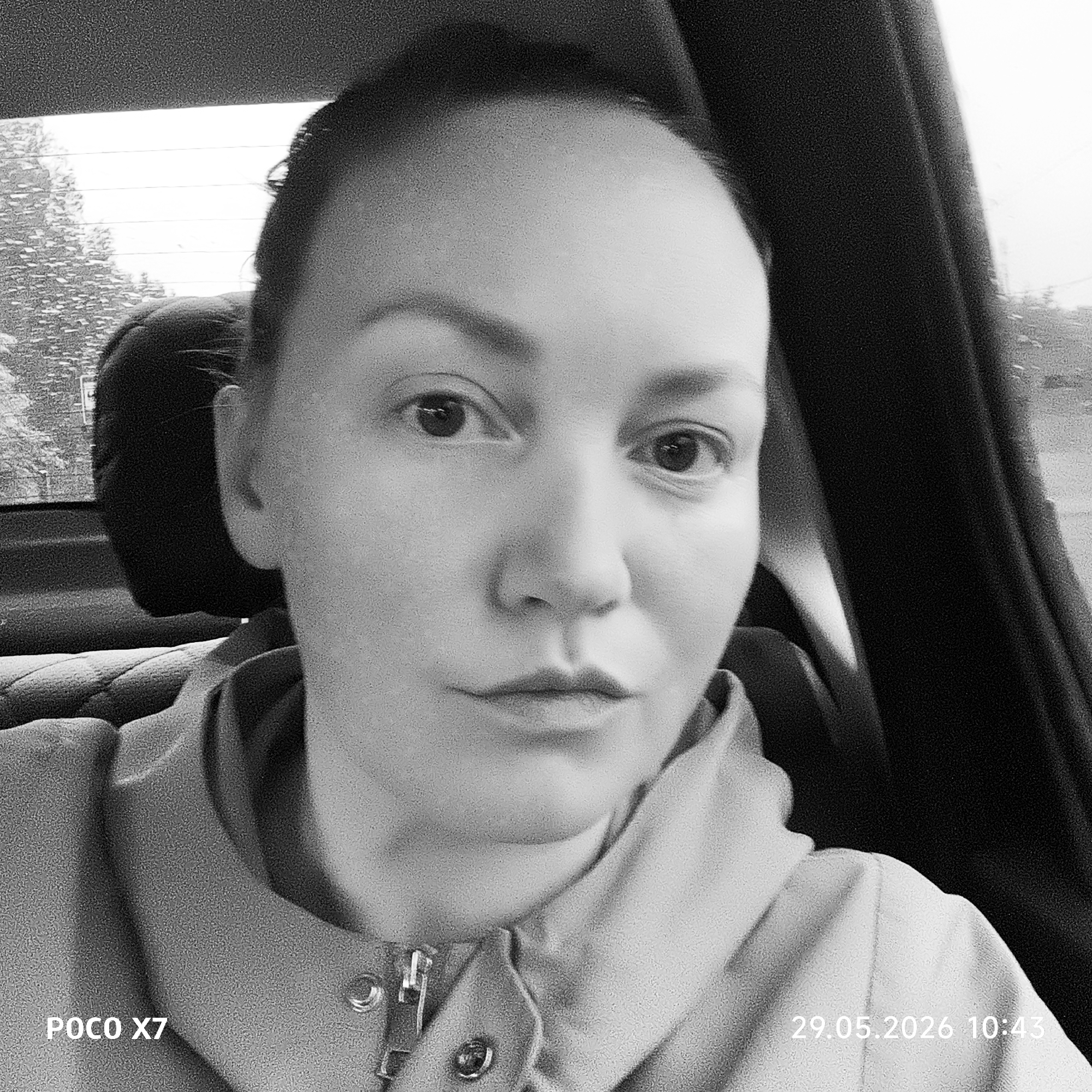 A close-up, black and white portrait of a person in a car, likely a selfie. The person appears to be female with closely cropped hair. She is wearing a jacket or hoodie. The background shows the interior of the car with a headrest visible behind her. Through the car window, trees are visible, likely outside of Chaikovsky, Russia. The bottom of the image contains the text "POCO X7" and "29.05.2026 10:43," indicating the phone model used to take the photo and the date and time it was taken.