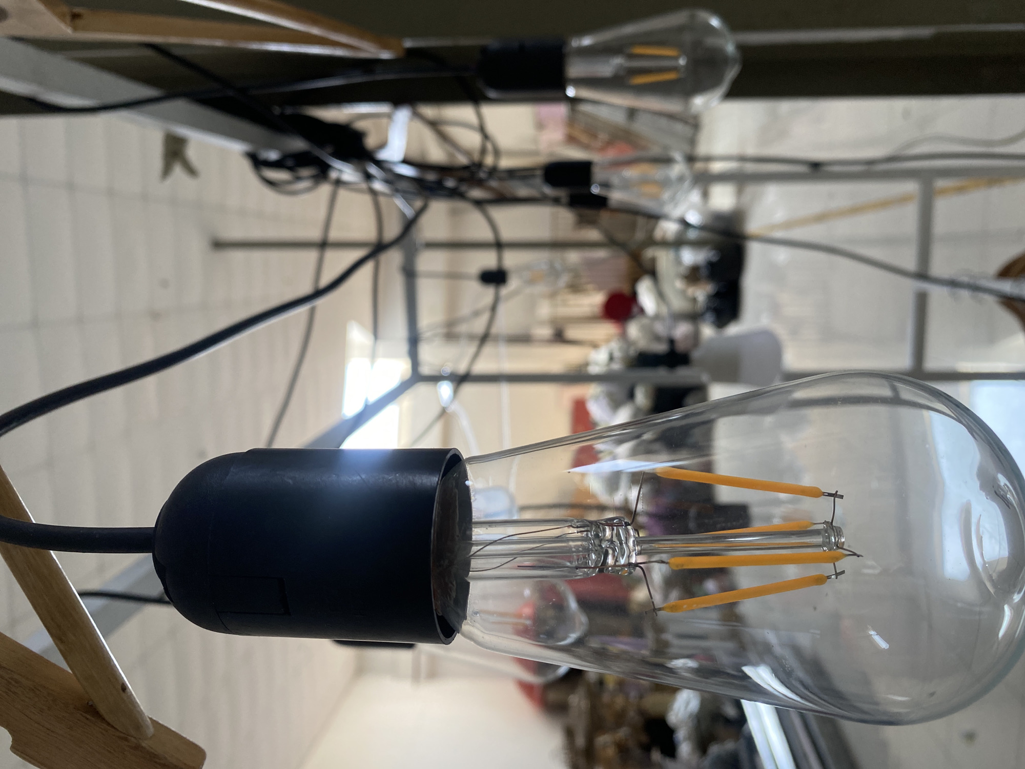 The image presents a close-up, upward-angled view of a modern Edison-style LED light bulb, currently unlit. The bulb features a clear glass envelope and houses distinct yellow-orange LED filament-like structures. It is securely screwed into a black electrical socket, from which a thick black wire extends away from the viewer. This primary light bulb is sharply in focus, dominating the foreground.
Behind the prominent bulb, several other identical LED light bulbs are visible, receding into the background and becoming progressively blurred due to a shallow depth of field. These bulbs, along with their associated black wiring, appear to be suspended from an overhead structure, possibly a shelf or framework made of dark metal or plastic. Numerous black electrical cables snake and crisscross throughout the mid-ground, suggesting a complex wiring setup. A wooden element, possibly part of a stand or furniture, is partially visible in the bottom left corner.
The setting appears to be an indoor environment, potentially a workshop, studio, or commercial space, indicated by the blurred light-colored walls and indistinct structural elements in the far background. There is no discernible activity taking place, and no people are visible. The image focuses solely on the lighting fixtures and their arrangement. No visible text is present in the frame. The location context provided is Davao City, Philippines.