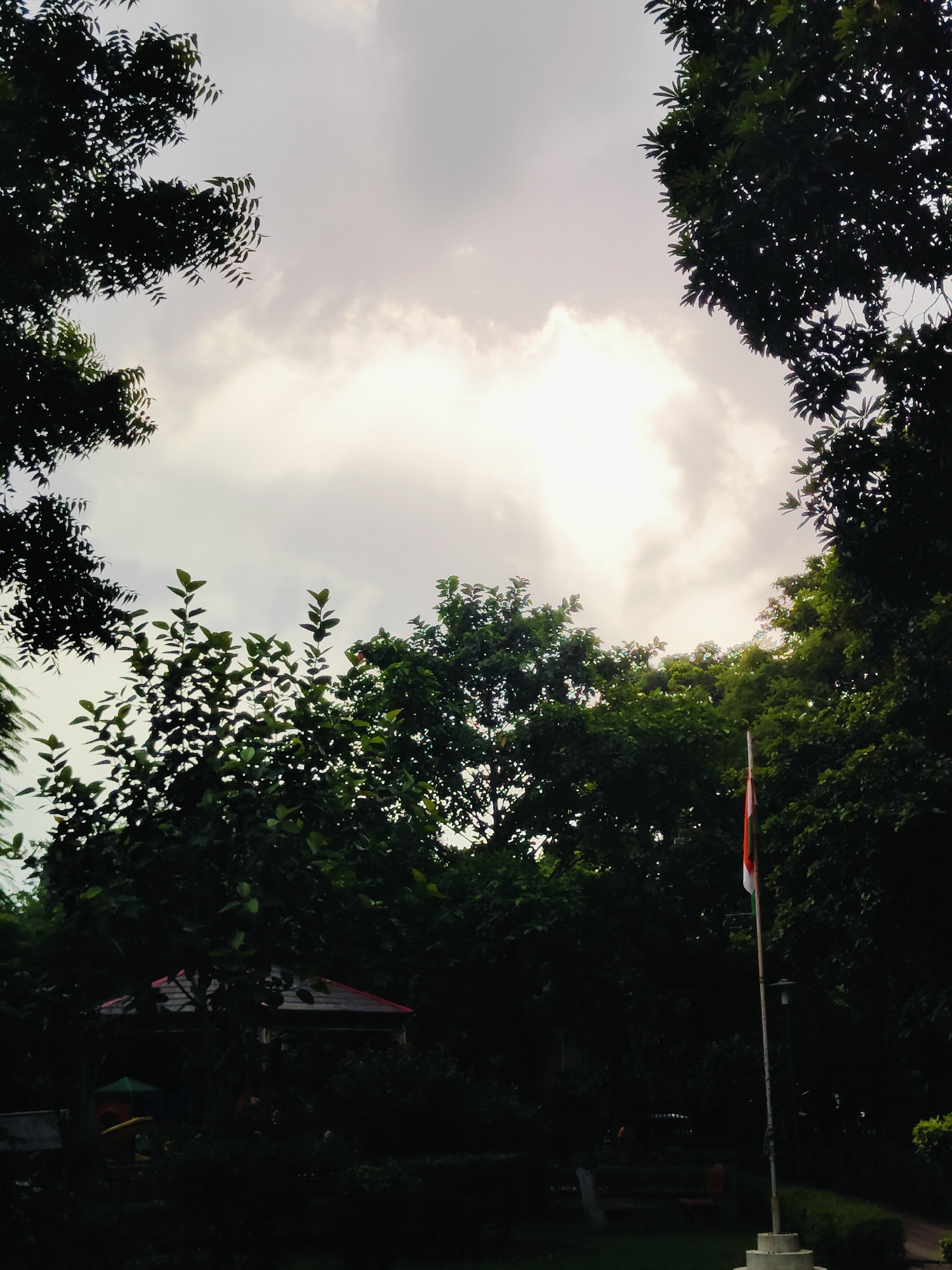 The image captures a view of the sky through dense foliage in Gurgaon, India. The sky is overcast with a bright, diffused light source, possibly the sun, breaking through the clouds. Silhouetted trees frame the sky from the top and sides, with varying degrees of darkness depending on their distance from the viewer. In the middle ground, a flag with three horizontal stripes of orange, white, and green is visible on a flagpole, suggesting a location of national significance. Below the flag, a streetlamp is partially visible. Further into the background, a structure with a red roof is discernible, partially obscured by the trees. The overall impression is of a serene, natural setting within an urban environment.