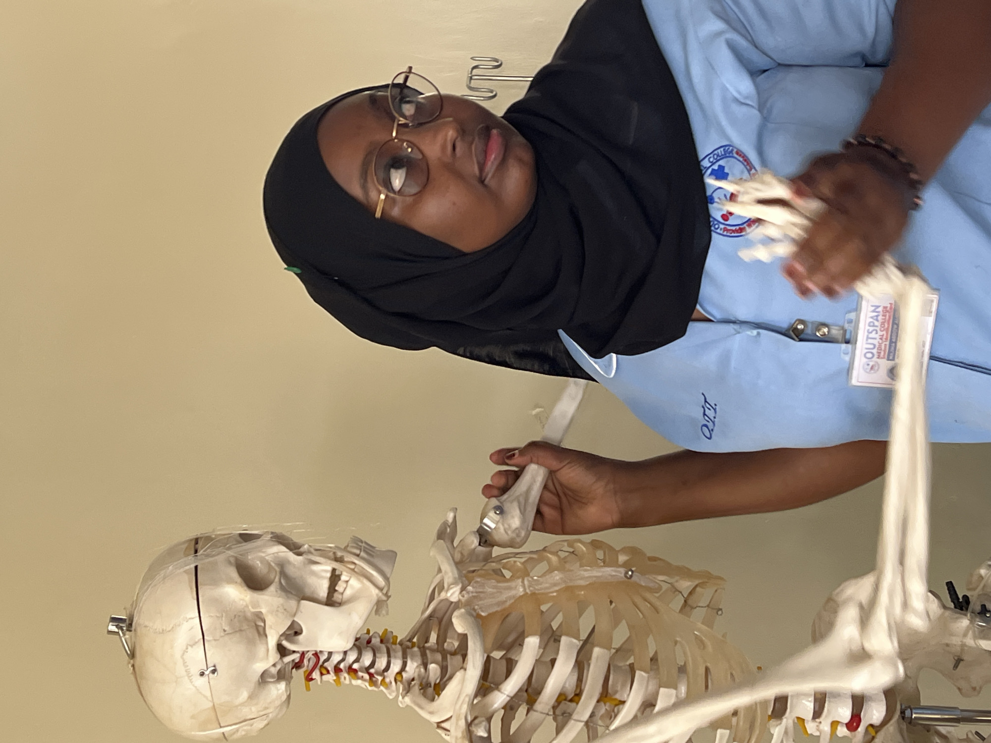 A dark-skinned young woman wearing a black hijab and a light blue medical scrub top with gold-rimmed glasses is standing to the right of a medical skeleton model. She is looking to her right, away from the camera. Her right hand is extended and touching the elbow joint of the skeleton. The skeleton is a life-size anatomical model, with its skull facing left and its vertebral column visible. A clear plastic sheet is draped over the top of the skeleton's skull. The background is a plain, light-colored wall. A name tag with "OUTSPAN MEDICAL COLLEGE" is attached to the woman's scrub top. The scene appears to be an educational or medical setting, possibly a classroom or laboratory, within Outspan Medical College in Nyeri, Kenya.