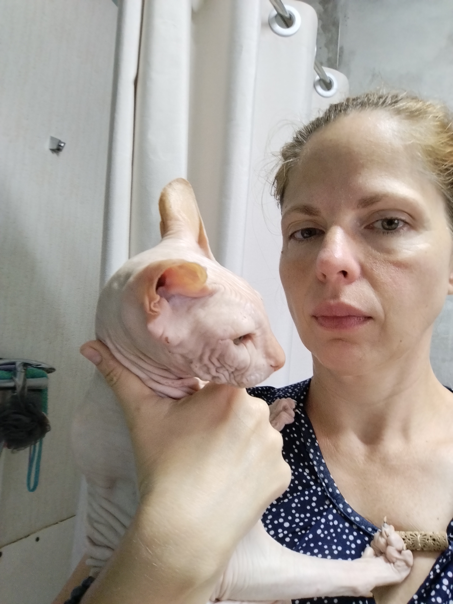 A woman is holding a hairless cat, likely a Sphynx breed, in her left arm. The cat is light pink and appears to be resting its head against her shoulder. The woman is looking towards the camera with a neutral expression. She is wearing a dark blue dress with white polka dots. In the background, there is a white shower curtain. The scene appears to be in a bathroom. There is no indication of the location being in Zavodoukovsk, Russia.