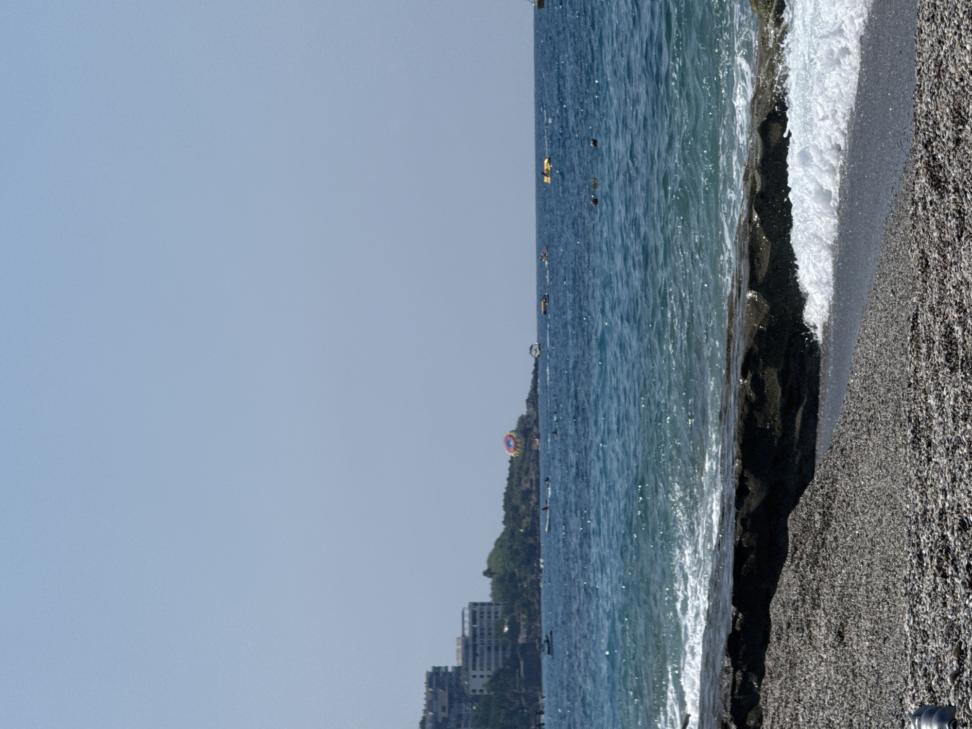A wide shot captures a pebble beach and a vibrant blue sea under a clear, light blue sky. In the foreground, the pebble beach slopes gently towards the water, with a dark, rocky outcrop breaking the shoreline. Waves crash onto the beach, creating white foam. In the middle ground, the sea is dotted with various watercraft including speedboats and yellow pedal boats. Farther out, a parasail with colorful stripes ascends into the sky. In the background, a line of buildings, some appearing to be hotels, are nestled on a green, hilly landscape. This scene is consistent with the coastal areas of Alanya, Turkey, known for its beaches and water activities.