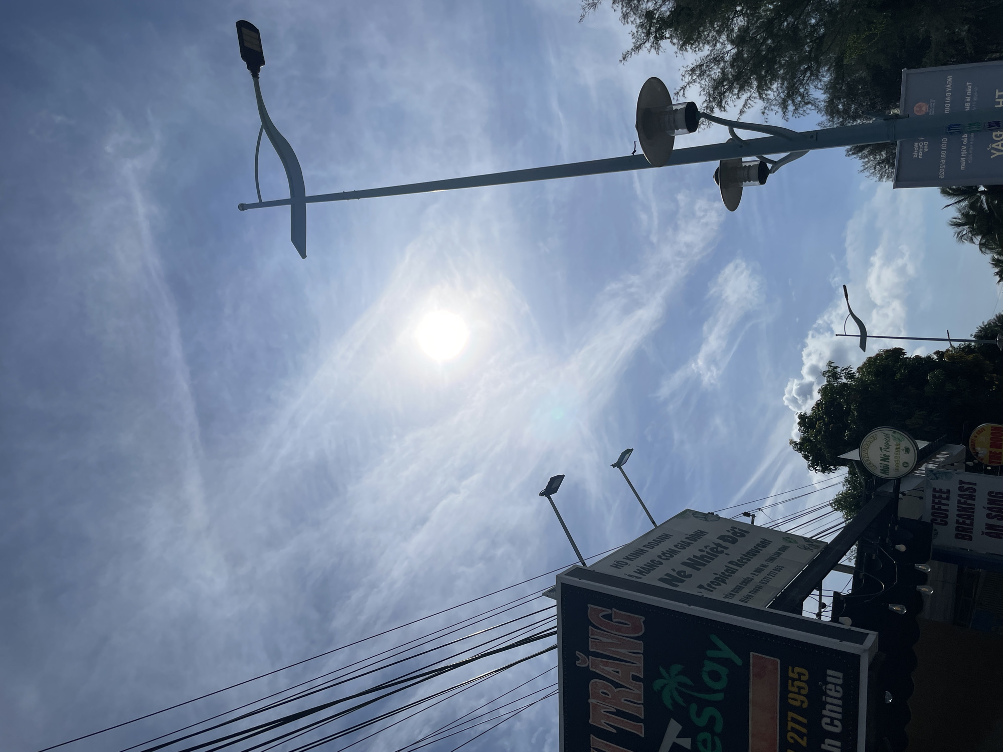 The image captures a street view in Phu Thuy Ward, Vietnam, under a bright, partly cloudy sky. Prominently featured are several streetlights and a cluster of power lines on the left side of the frame. A sign for "Né Nhiệt Đới Tropical Restaurant" is visible, along with other signs displaying Vietnamese text and phone numbers, suggesting a commercial area. Trees and foliage are present in the background, indicating an outdoor setting. The sun is visible and positioned towards the upper center of the image, casting a glare. The composition is dominated by the sky and urban infrastructure.