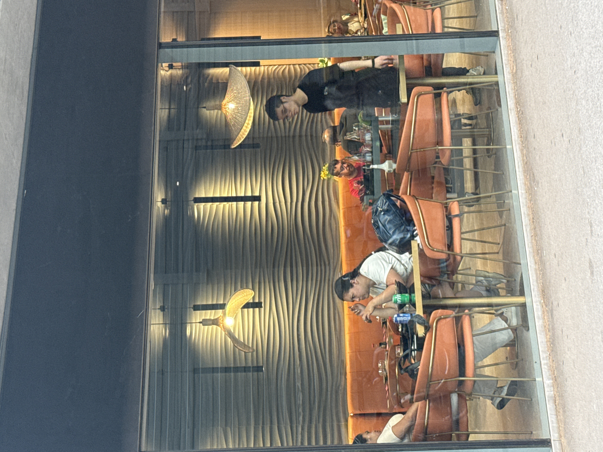 This is a medium-shot, eye-level, outdoor-view photograph of a restaurant interior, seen through a large glass window. The lighting suggests it is daytime. The restaurant has a modern aesthetic with orange upholstered booth seating and chairs with thin, gold-colored legs. The back wall features a textured, wavy pattern in muted tones, with two large, pendant light fixtures with woven shades hanging from the ceiling.
In the foreground, a woman with dark hair pulled back is seated at a table, looking down with her hands clasped in front of her. She is wearing a white t-shirt. To her left, another person, partially visible, is also seated. On the table are various items including a blue can, a green can of Sprite, a bowl of food, and a bottle of soy sauce.
Further into the restaurant, a young man wearing glasses and a black t-shirt is standing and looking to his left. He appears to be a staff member. Behind him, other patrons are seated at tables, including a man in a red shirt and another man with a shaved head. The reflections on the glass window are noticeable, obscuring some of the details within the restaurant.
The exterior of the building is not clearly visible, but the presence of a sidewalk in the foreground indicates an urban setting. The overall impression is of a contemporary dining establishment.