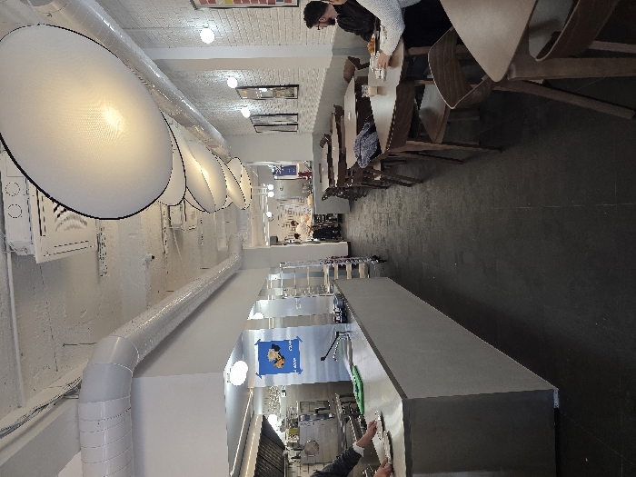 This image captures the interior of a "4 Good Food" restaurant, identifiable by a blue "4 GOOD FOOD - GRAB" poster on the rear wall, located in Sofia, Bulgaria. The long, narrow establishment is divided into a service/kitchen area on the left and a dining area on the right, separated by an aisle.
On the left, a long, light grey counter with a stainless steel top and an integrated sink runs along the wall. A person in a dark jacket is positioned behind this counter, holding a plate. Behind the counter, a professional kitchen with stainless steel appliances, including a large ventilation hood, is visible, with at least one additional staff member working further back. A multi-tiered stainless steel rolling cart is located near the counter.
The dining area on the right features multiple square dark wooden tables paired with matching wooden chairs, arranged along a white brick-textured wall. Framed artwork is hung on this wall. Two individuals are seated at a table in the mid-ground: one person in a dark top is oriented away from the camera, and another in a light-colored sweater faces towards the camera, obscured. A hot beverage cup and a plate with food are on their table. A blue jacket is draped over a chair at an unoccupied table nearby.
The ceiling features exposed white ventilation ducts and a series of large, elliptical white light fixtures suspended at various heights, providing ambient illumination. Additional spherical wall-mounted lights and recessed ceiling lights are present. The floor throughout the establishment is dark grey.