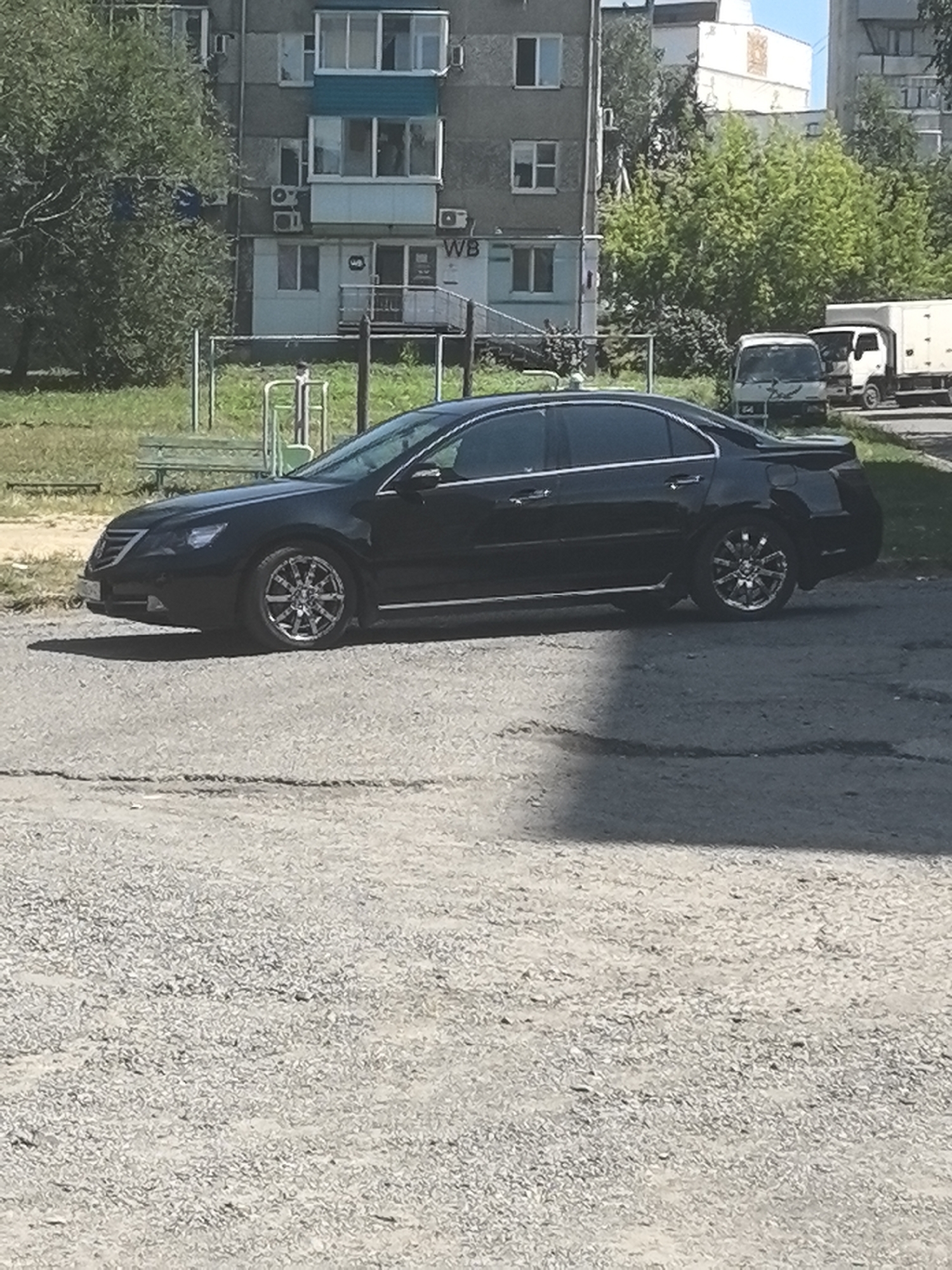 A black sedan is parked on a cracked asphalt surface. The car is facing left and is partially obscured by a shadow. In the background, there is a building with multiple windows and balconies, and a playground with a green bench. Trees and parked vehicles, including a white truck, are also visible in the distance. The overall scene suggests an urban environment, possibly a residential area.