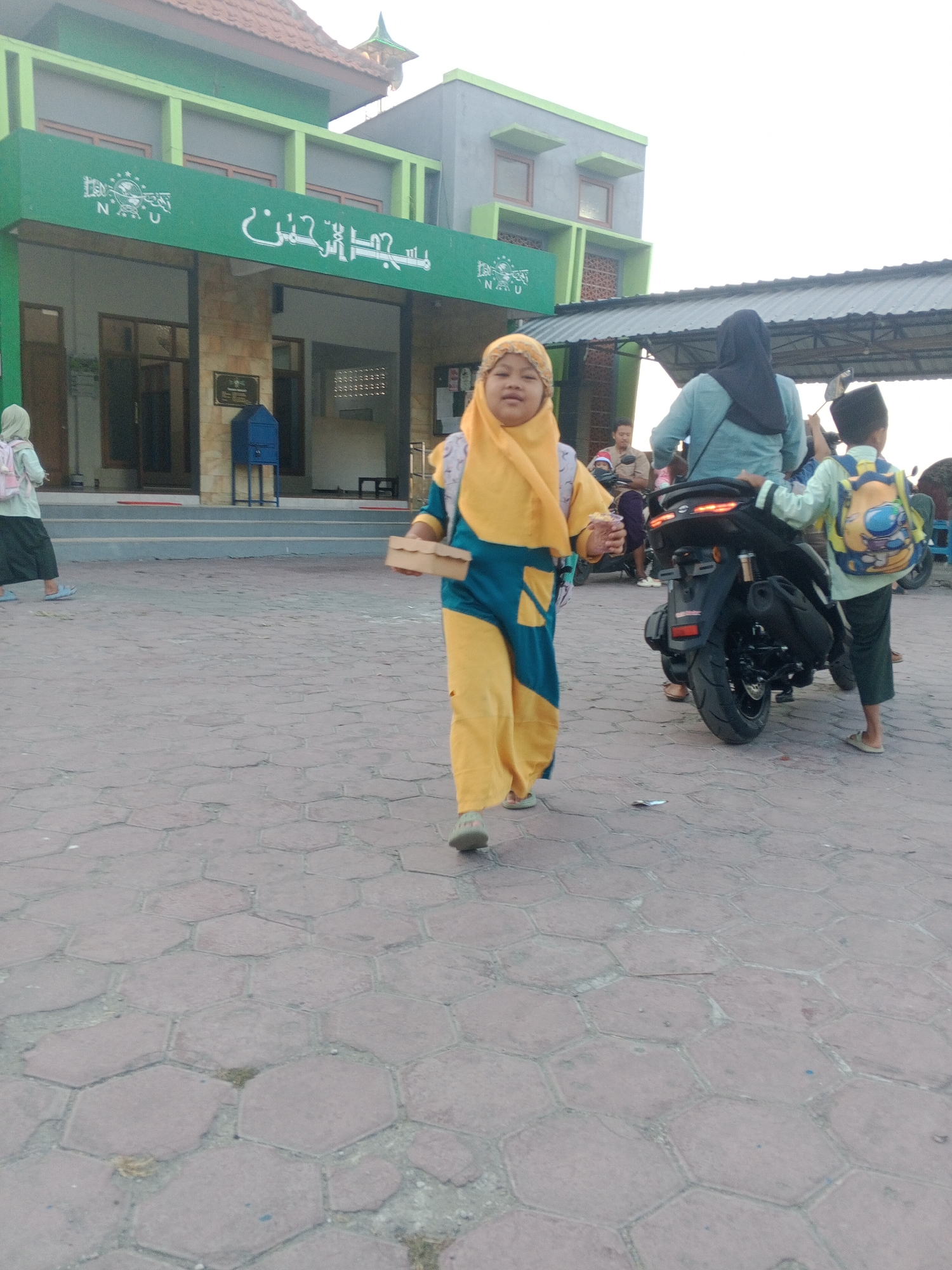 The image depicts an outdoor scene in front of a building with green trim and Arabic calligraphy. A young girl wearing a yellow hijab and a teal and yellow outfit is walking towards the viewer, carrying a small brown box. To her right, a black motorcycle is parked. On the motorcycle, a person wearing a blue headscarf is seated, and a young boy wearing a light blue shirt and a yellow backpack stands next to it. Another child in a hijab and backpack is visible in the background to the left. The ground is paved with reddish-brown hexagonal tiles. The building appears to be a place of worship or a school, possibly located in Lembah, Indonesia, given the presence of Arabic script. The sky is overcast.