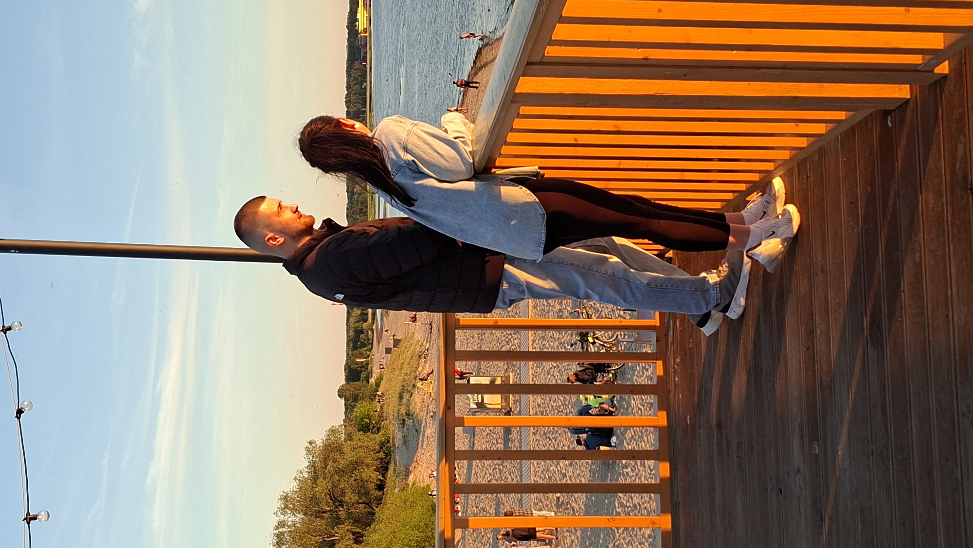 Two people, a man and a woman, are standing on a wooden pier overlooking a beach and the sea. The man is wearing a black puffer jacket and light blue jeans. The woman is wearing a light blue denim jacket over black leggings. They are facing each other, with the woman leaning on a wooden railing. The scene is bathed in the warm, golden light of sunset, indicating it is late evening. The sky is clear with some wispy clouds. The beach is populated with a few other people and some parked bicycles. The sea is calm with a few individuals in it. The wooden pier has a slatted overhead structure creating a striped shadow effect on the surface. The overall mood appears peaceful and romantic. The location is identified as Tallinn, Estonia.