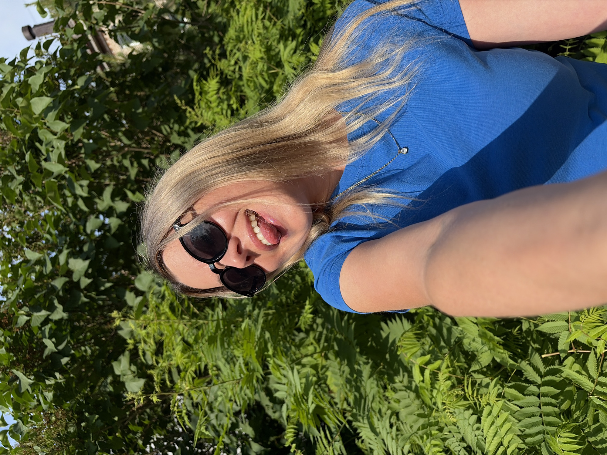 A young, fair-skinned woman with blonde hair is pictured outdoors, in front of lush green foliage, likely a park or garden in Chaikovsky, Russia. She is wearing a bright blue t-shirt and black sunglasses, and her tongue is sticking out playfully. The sunlight casts dappled shadows on her and the surroundings, suggesting a sunny day. She is holding a phone or camera out of frame, taking a selfie.