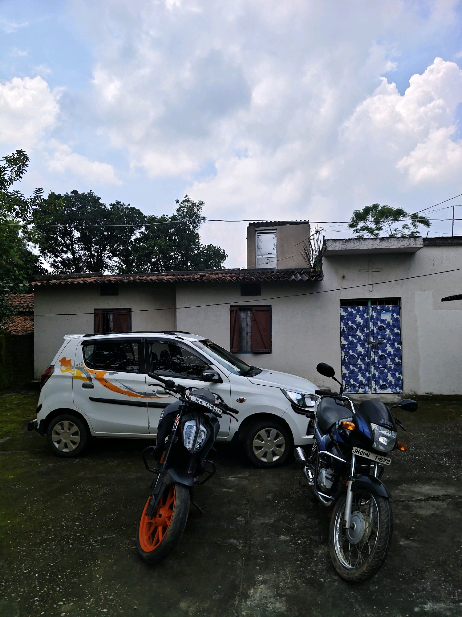 In Sosai, India, under a cloudy sky, two motorcycles and a white hatchback are parked on a paved surface in front of a light-colored building. The building has a tiled roof and features brown wooden shutters on some windows, and a pair of blue floral patterned doors on another. A cross emblem is visible above the patterned doors. The white car has an orange and yellow graphic design on its side. The motorcycle in the foreground is black with orange wheels and has a license plate reading "JH 01 ES 8829". The second motorcycle is dark blue and black, with a license plate reading "JH 01 N 1872". Trees surround the building, and power lines are strung overhead.