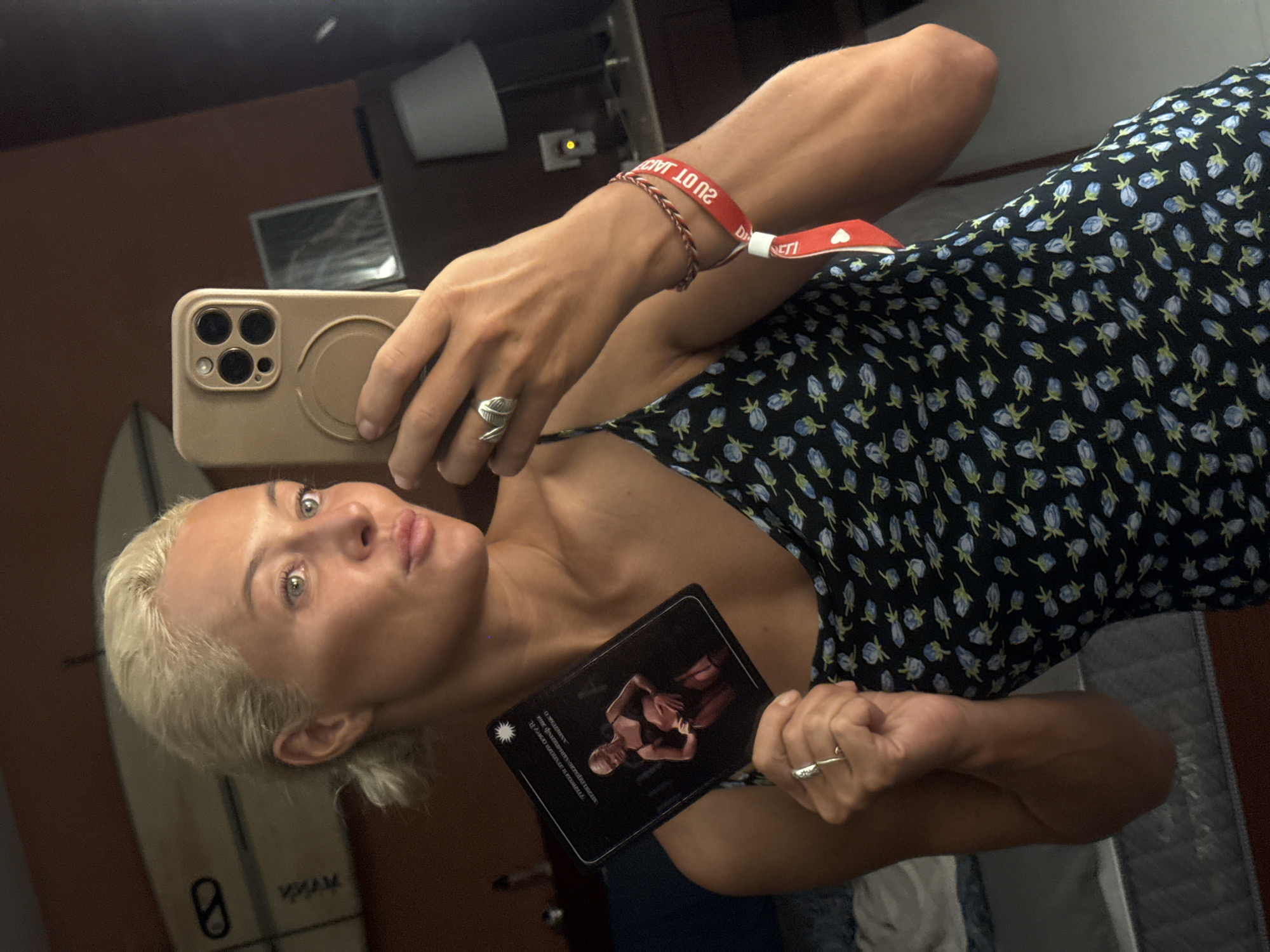 The media file is a vertical, slightly angled selfie taken in what appears to be an indoor setting with warm-toned walls and reflections in a mirror. The primary subject is a fair-skinned woman with short, blonde hair and blue eyes. She is holding a smartphone in her right hand, which is capturing her reflection.
She is wearing a dark, floral-patterned dress with small blue flowers. On her left wrist, she has a red festival-style wristband with text that appears to read "202102" and "DI...NETI". She is also wearing a silver ring on her left ring finger.
In her left hand, she holds a black card with an image of two people in what looks like an intimate pose. The card has some text in a light color, but it is mostly illegible. Her facial expression is one of playful puckering of the lips.
The background shows reflections of various objects, including a surfboard standing upright, a piece of furniture that might be a bed or couch with grey coverings, and some electronic equipment on the wall. The lighting suggests it could be daytime or well-lit indoors. The overall mood conveyed is casual and playful.