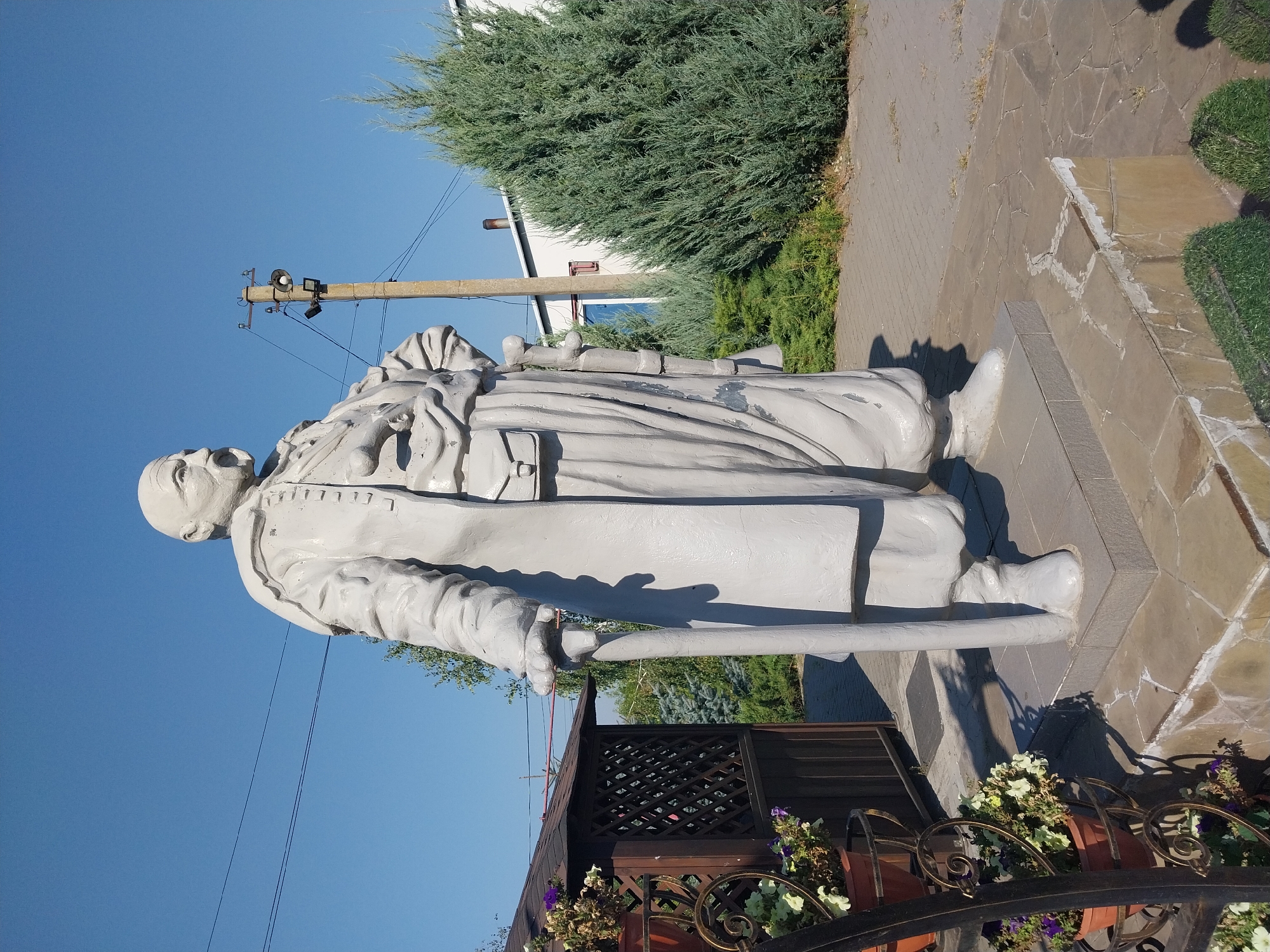 The image shows a life-sized, gray stone statue of a man in historical attire, standing on a stone pedestal in Petrykivka, Ukraine. The man has a stern expression, a mustache, and is wearing a long coat, a sash with pouches, and boots. He is holding a cane in his left hand. Behind the statue, there is a small wooden gazebo with potted flowers, a tall utility pole with electrical wires, a green conifer tree, and a white building. The sky is clear and blue, indicating a sunny day. The ground around the statue is paved with stone tiles and there are also some green bushes and grass. The statue is positioned outdoors, likely in a public space or park.
