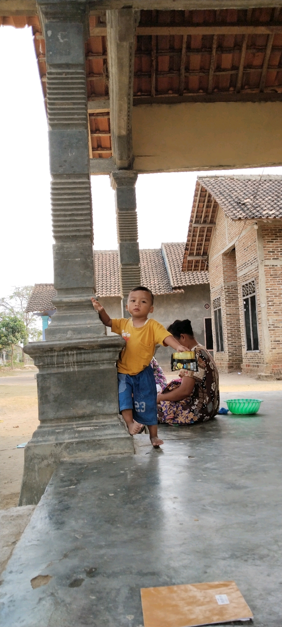The image depicts a child standing on a concrete porch in front of a building in Tanjung Ratu Ilir, Indonesia. The child, wearing a yellow t-shirt and denim shorts, is positioned to the left of a large, decorative concrete pillar. Behind the child, a woman is seated on the porch, wearing a patterned dress. To the right of the woman, a green plastic bowl is visible. The building behind them appears to be constructed of brick with a tiled roof. The overall environment suggests a rural or semi-rural setting.