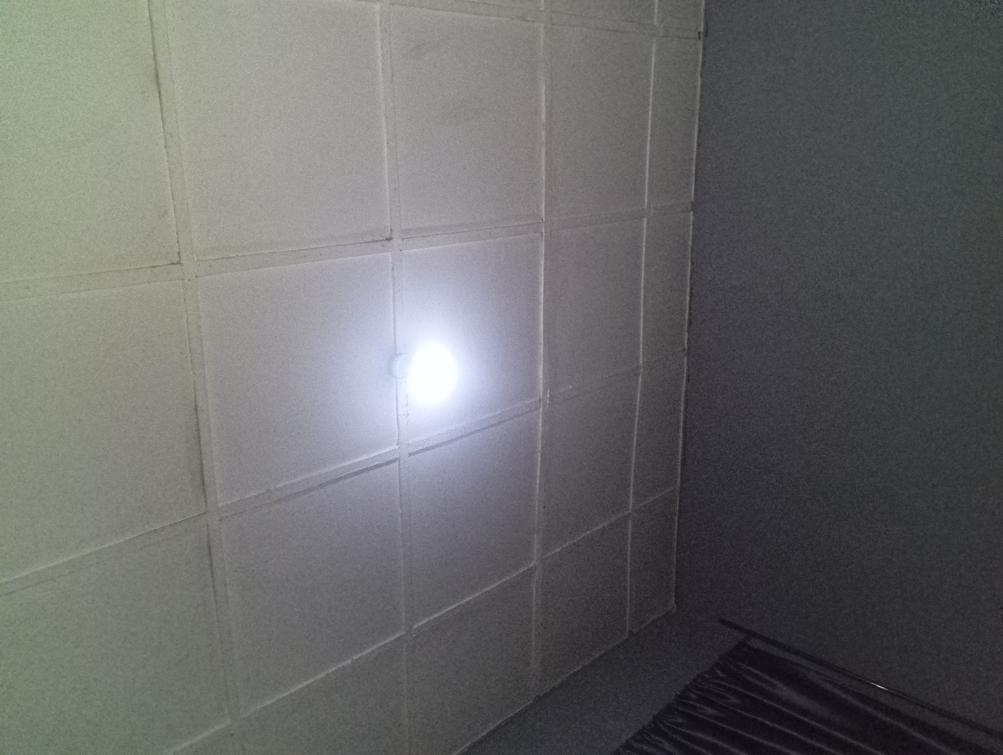 An indoor, eye-level, medium shot photograph captures a ceiling with a grid of square panels, illuminated by a bright, overhead light. The ceiling tiles are textured and light gray, with darker grout lines forming the grid. The lower portion of the image shows a dark gray wall with a section of black curtains on the left. The scene appears to be in a room with no people present. The location is unknown.