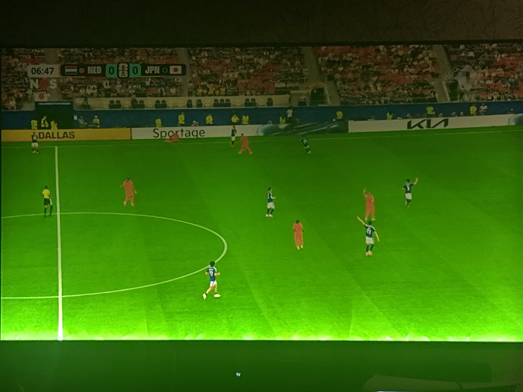 The image shows a digital display of a soccer match. The score is NED 0 - JPN 0, and the time is 06:47. There are several players on the green field, wearing orange and blue/white uniforms. The background shows stadium seating filled with spectators, and advertisements along the wall, including "DALLAS", "Sportage", and "KIA". The image is highly pixelated and has a green tint, possibly due to lighting conditions or the display technology. There is no discernible location in the city of Leiden, Netherlands visible in the image; it appears to be a generic stadium setting for a soccer game.