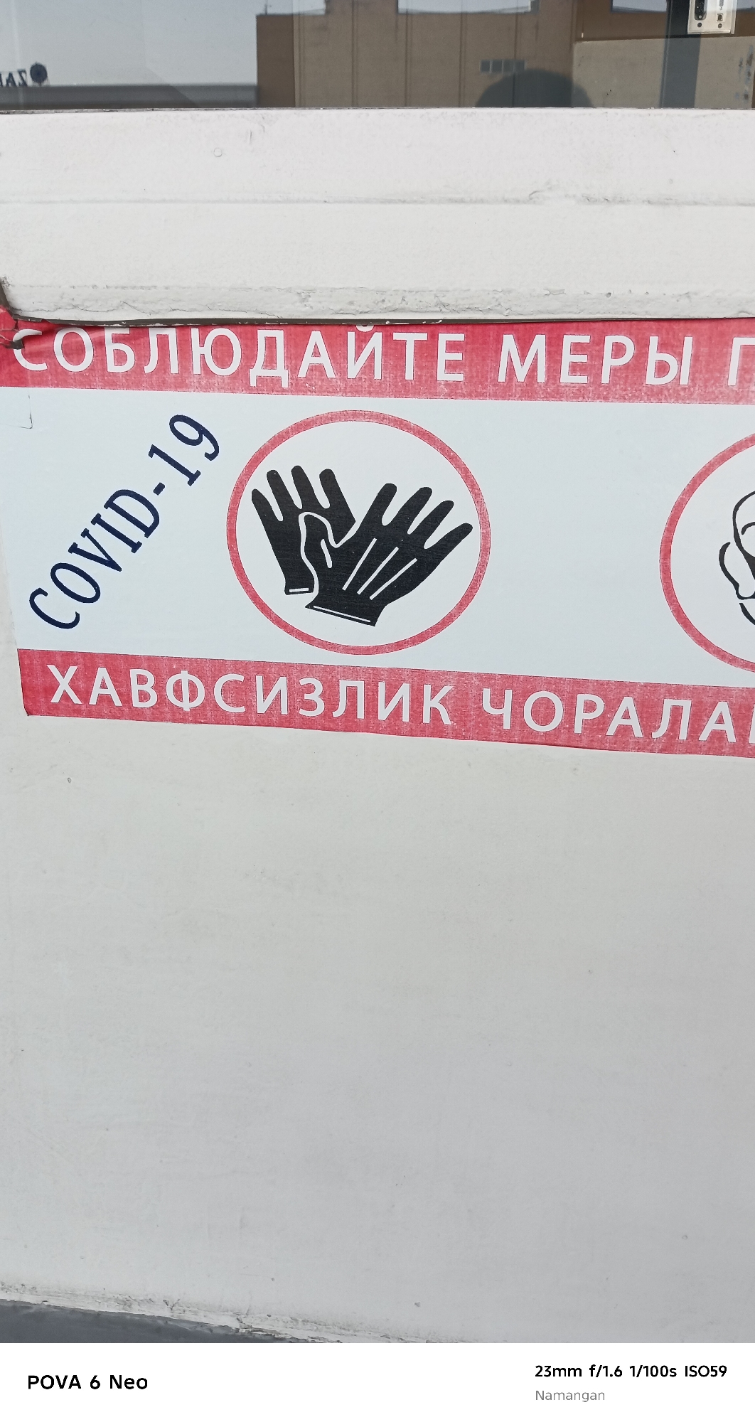 The image shows a close-up of a sign on a white wall. The sign has red and white lettering and graphics. The top line of text, in white capital letters on a red background, reads "СОБЛЮДАЙТЕ МЕРЫ". Below this, a curved blue text reads "COVID-19". To the right of the COVID-19 text is a graphic of two black gloved hands inside a red circle. Below the COVID-19 text and the gloved hands graphic is another line of text in white capital letters on a red background which reads "ХАВФСИЗЛИК ЧОРАЛА". The bottom of the image shows text indicating "POVA 6 Neo" and camera settings "23mm f/1.6 1/100s ISO59 Namangan". The overall impression is of a public health warning related to COVID-19, likely posted in a public space within Namangan City, Uzbekistan.