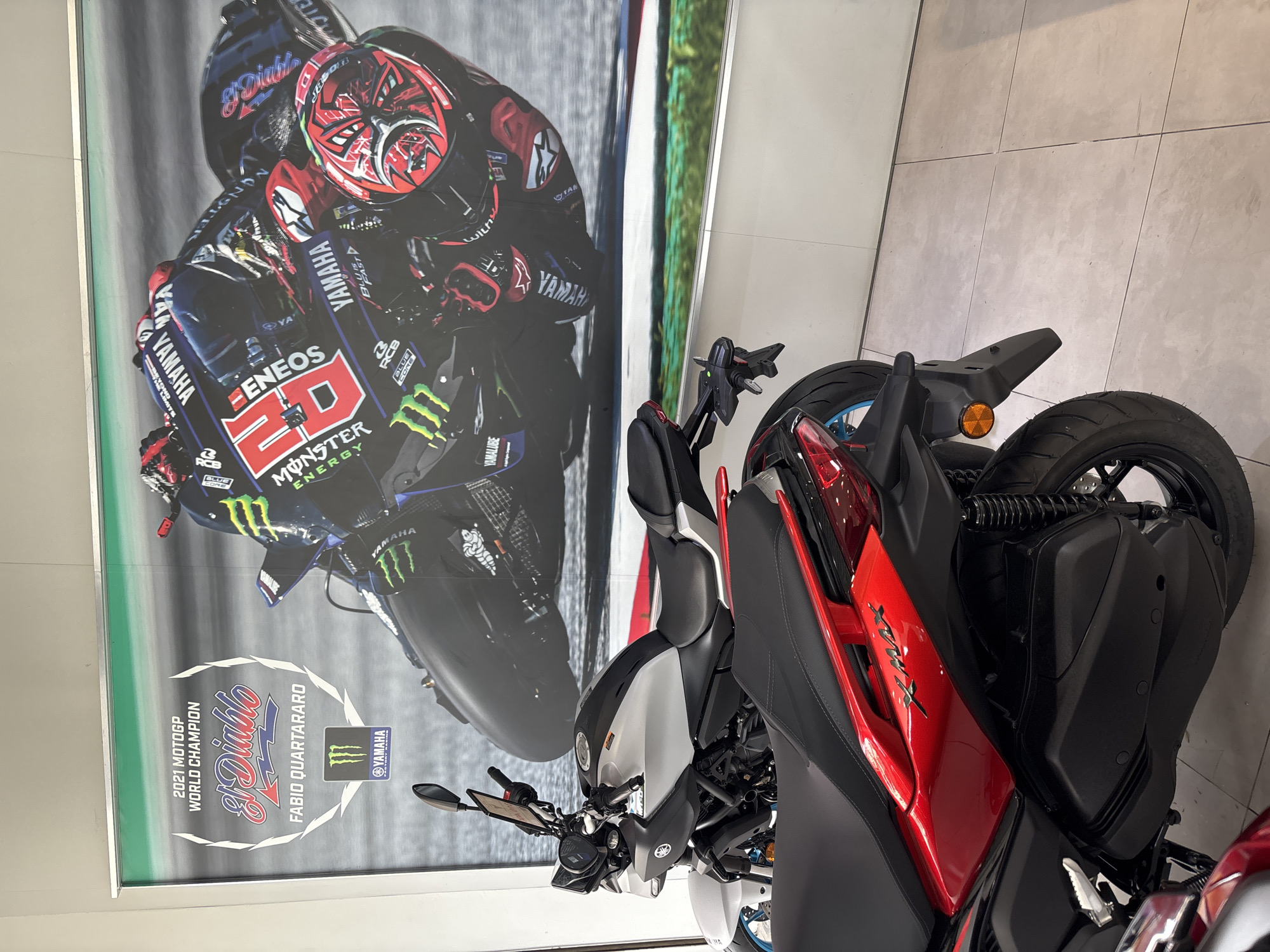 The image shows a Yamaha XMAX scooter parked indoors, in front of a poster of Fabio Quartararo's 2021 MotoGP World Champion Yamaha motorcycle. The poster is vertically oriented and features the Yamaha branding and sponsors, including ENEOS and Monster Energy. The scooter is red, white, and black, with its rear facing the viewer. The setting appears to be a dealership, possibly in Baan Bo Phut, Thailand.