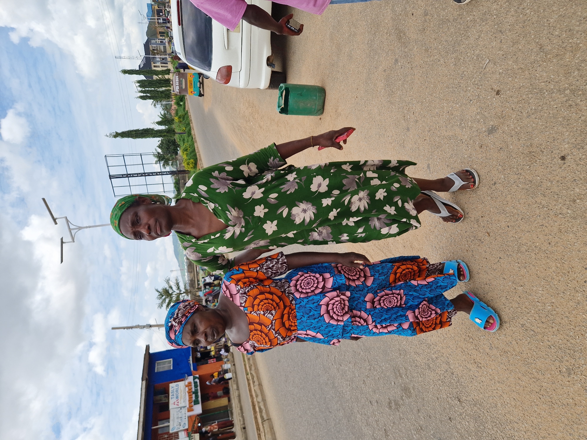 Two women walk along a paved road in Kabong, Nigeria. The woman on the left wears a blue and pink patterned dress with a matching headscarf. She has blue flip-flops on her feet. The woman on the right wears a green floral dress and a green headscarf. She wears white flip-flops. In the background, there are buildings, trees, a white car, a tuk-tuk, and streetlights. A green plastic container sits on the side of the road. The sky is cloudy.
