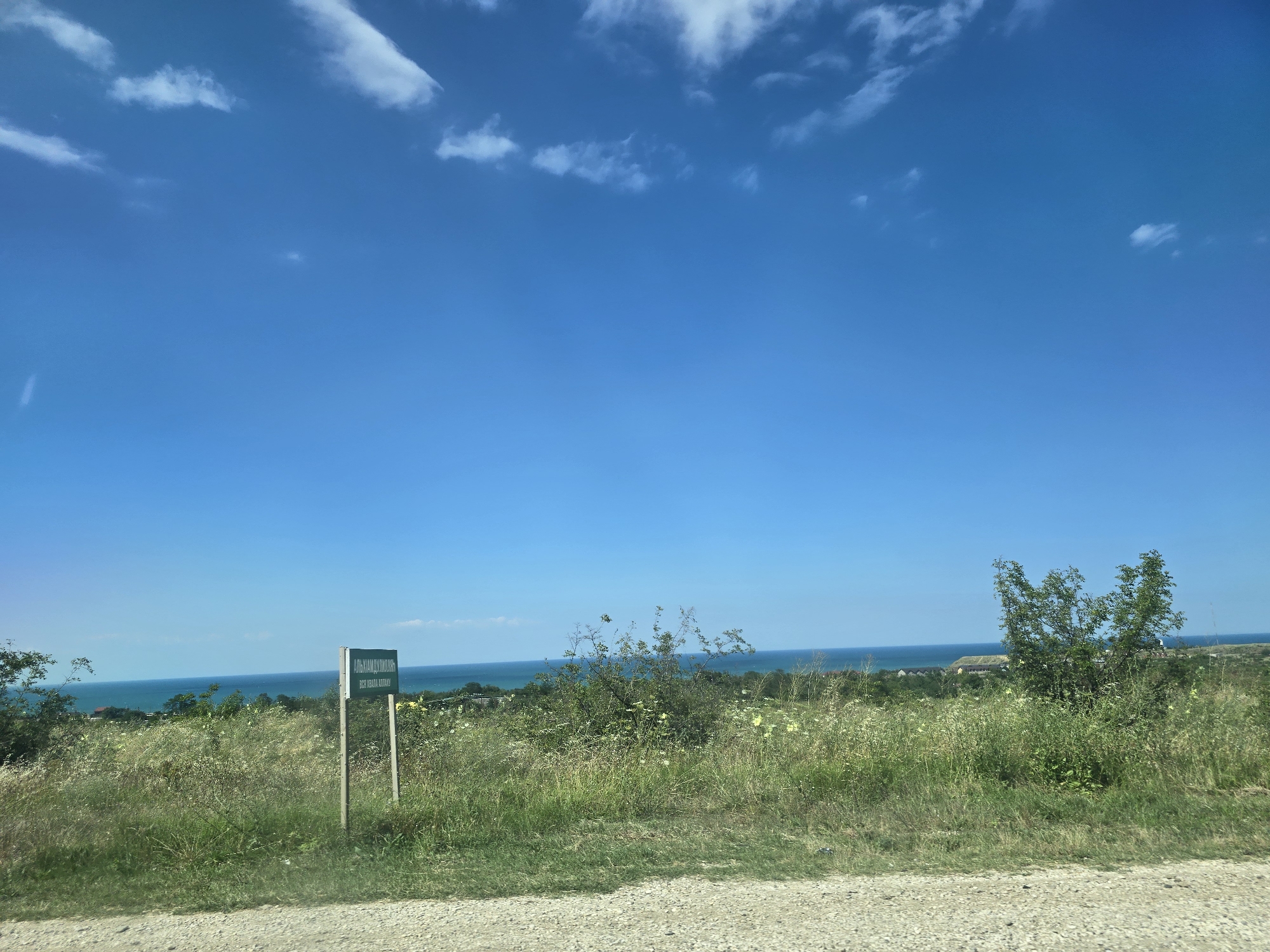 The image shows a wide, sunlit view of a grassy hillside sloping down towards a body of water, likely the sea, under a clear blue sky with scattered white clouds. In the foreground, a dirt road or path is visible. A green sign with white lettering stands on a wooden post in the middle ground. The sign is in Russian and appears to be a directional or informational marker. The hillside is covered in tall, green grass and wildflowers, with some scattered bushes and a small tree on the right side of the frame. In the distance, there are more trees and some buildings, suggesting a settlement. The overall impression is one of a natural, rural landscape on a bright, clear day. The provided crops confirm the presence of the sky, the sign, and the natural environment. The location is identified as Ullubiiaul, Russia.