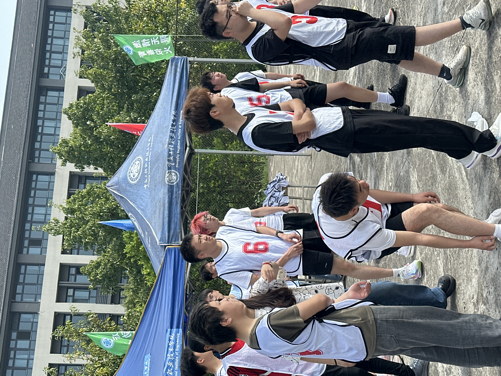 This outdoor image captures a gathering of young men, likely students, under a bright, clear sky on a sunny day. The scene takes place on a concrete ground, possibly a sports court or campus area, with a modern multi-story building featuring many windows and lush green trees in the background.
The subjects are dressed in sportswear, predominantly white sleeveless vests with red numbers (such as '5', '6', '10') worn over black t-shirts, paired with either black shorts or pants. Some individuals are standing attentively, while others are seated or resting. One young man is notable for his pink-dyed hair. Their expressions appear calm and relaxed, suggesting a break or a moment of waiting during an event.
In the foreground and midground, two blue tents or canopies are set up. Both display the Chinese text "重庆工程职业技术学院" (Chongqing Vocational Institute of Engineering) along with its English translation. Several flags are also visible; a green flag prominently features the Chinese phrases "青春炒火" (Youth Stir-fried Fire) and "燃計天則" (Burn the Law of Heaven), indicative of event slogans, and a red flag is also present. The attire of the participants and the presence of labeled tents and flags suggest a student event, possibly a sports competition or an organizational gathering, taking place at the Chongqing Vocational Institute of Engineering in Jiangjin, China.