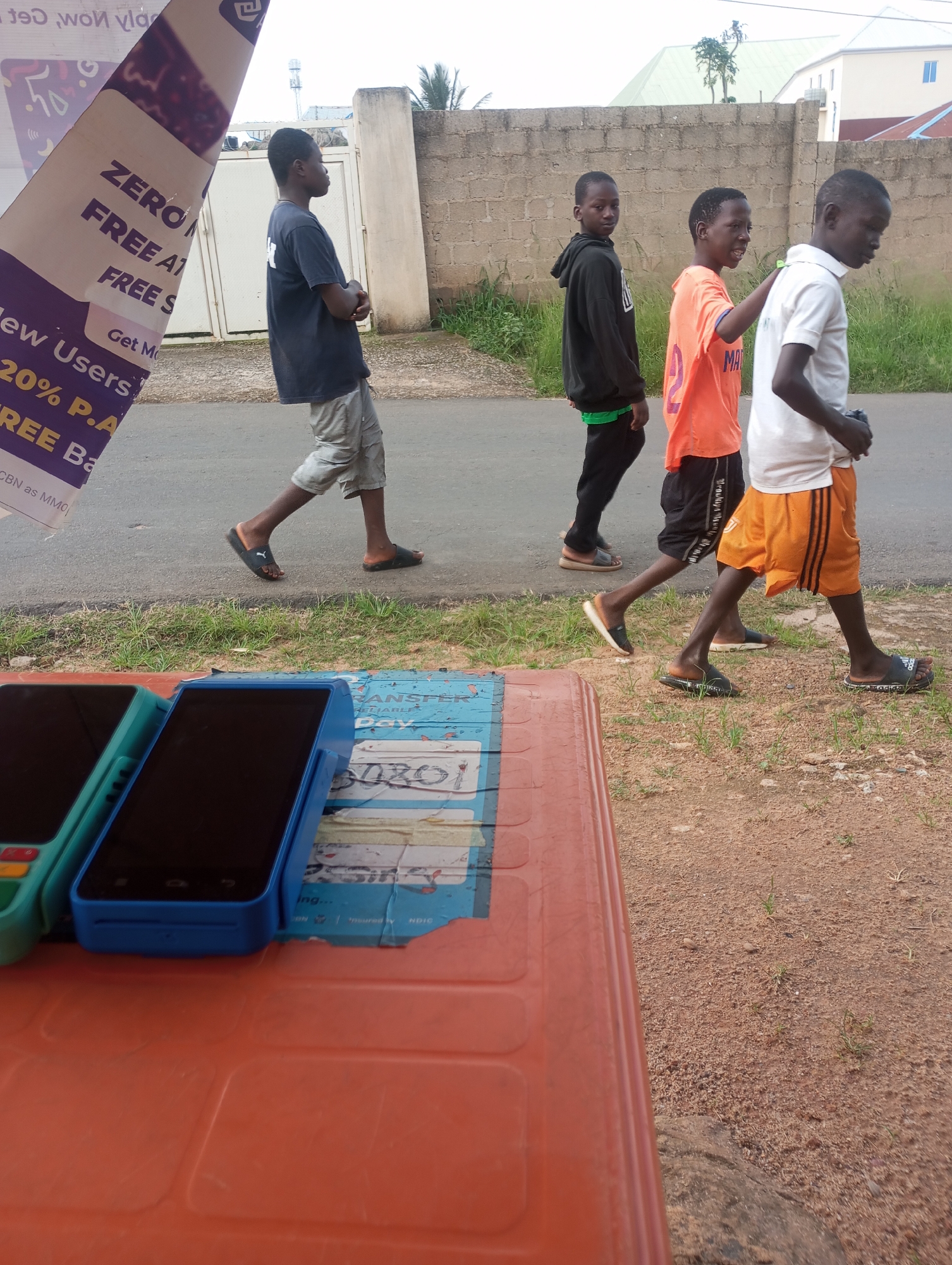 Four young males are walking on a paved road with a sandy shoulder in Jos, Nigeria. In the foreground, two POS (point of sale) terminals are visible on an orange plastic surface. The first POS terminal is teal, and the second is blue. The males are walking from left to right. The first male, closest to the foreground, is wearing a dark blue t-shirt and light grey shorts. The second male wears a black hoodie over a green shirt and black pants. The third male is wearing a bright orange t-shirt and black shorts. The fourth male, on the far right, is wearing a white t-shirt and orange shorts with black stripes. They are walking past a low brick wall and some vegetation. A large, angled banner with text is on the far left of the image. The sky is overcast.