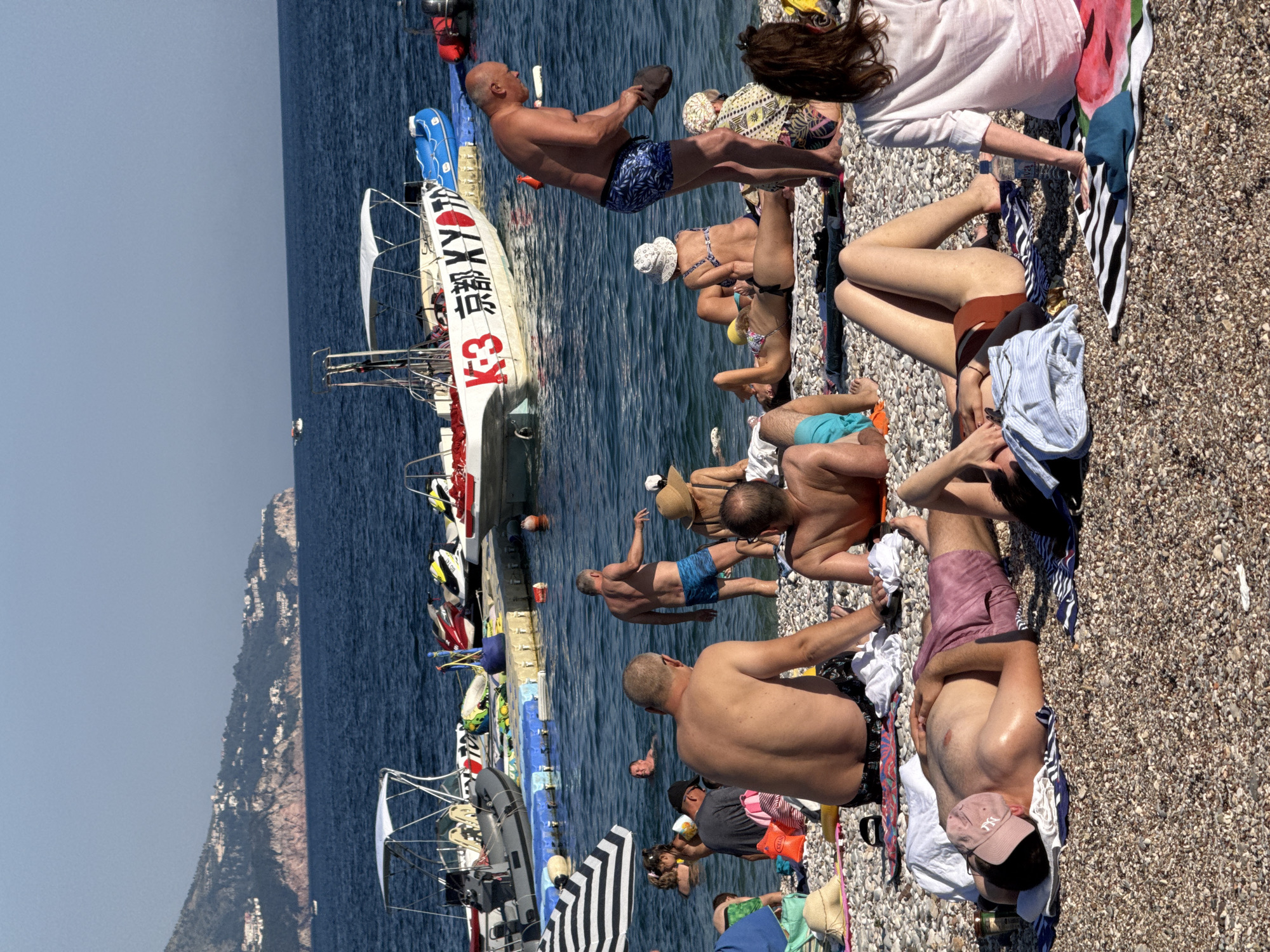 The image depicts a crowded beach in Budva, Montenegro, with numerous people sunbathing on a pebble shore and swimming in the clear blue sea. Several boats, including speedboats and a larger vessel with "K-3 京都 XY" written on its side, are anchored near a floating dock. Beach umbrellas and towels are scattered across the sand. The background shows a mountainous coastline dotted with buildings. The scene is bathed in bright sunlight, suggesting a warm, sunny day.