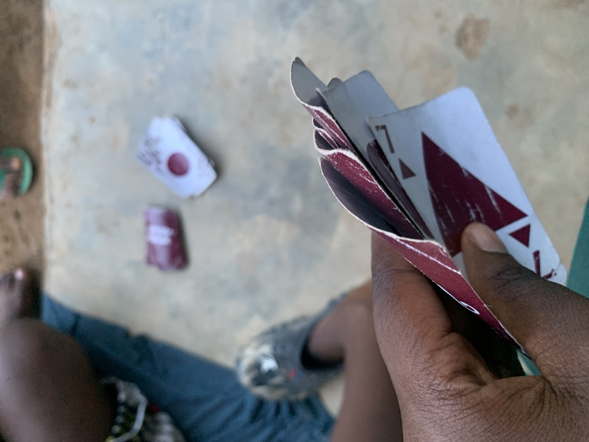 The image, captured in Keffi, Nigeria, presents an overhead view of a person engaged in a card game outdoors during the daytime. The primary subject is a dark-skinned hand holding a fanned-out hand of playing cards, with the maroon and white backs of the cards prominently displayed. One card in the hand clearly shows the number "7" in the top left corner, along with a prominent red, triangular or chevron-shaped suit symbol.
In the blurred background, parts of a person's legs and feet are visible, indicating they are seated on the ground. One foot is bare, while the other wears a grey slipper. Several discarded playing cards are scattered on the light-colored ground, which appears to be concrete or packed earth. One discarded card shows a distinct round, red design with some lighter, indistinct markings around it, likely part of its back pattern. The scene conveys a moment of casual recreation under natural light.
