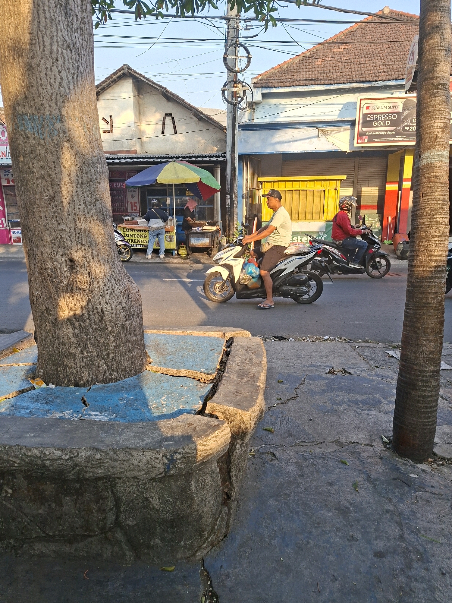 In Surabaya, Indonesia, a street scene unfolds at street level under a clear sky. A large tree trunk dominates the left foreground, its rough bark textured and casting dappled shadows. In front of the tree, a cracked concrete planter box, painted in patches of blue and gray, shows signs of wear and tear.
To the right, a paved street runs horizontally across the image. Two men on motorcycles are captured in motion. The man on the left rides a white scooter, wearing a light-colored t-shirt, dark shorts, and flip-flops, with a plastic bag hanging from his handlebars. The man on the right, wearing a red jacket and helmet, rides a black scooter.
In the middle ground, a street-side vendor stall is set up with a colorful umbrella providing shade. Behind the stall, a building with a brown tiled roof and a light blue facade stands. A prominent advertisement for "DIARUM SUPER ESPRESSO GOLD" is displayed on the building's exterior. Yellow metal gates and red and yellow striped vertical elements are visible on the building's lower level. The street is lined with power lines and poles, indicating an urban environment. The ground is paved with asphalt, showing cracks and debris in some areas. The overall impression is a slice of everyday life in a bustling Indonesian city.