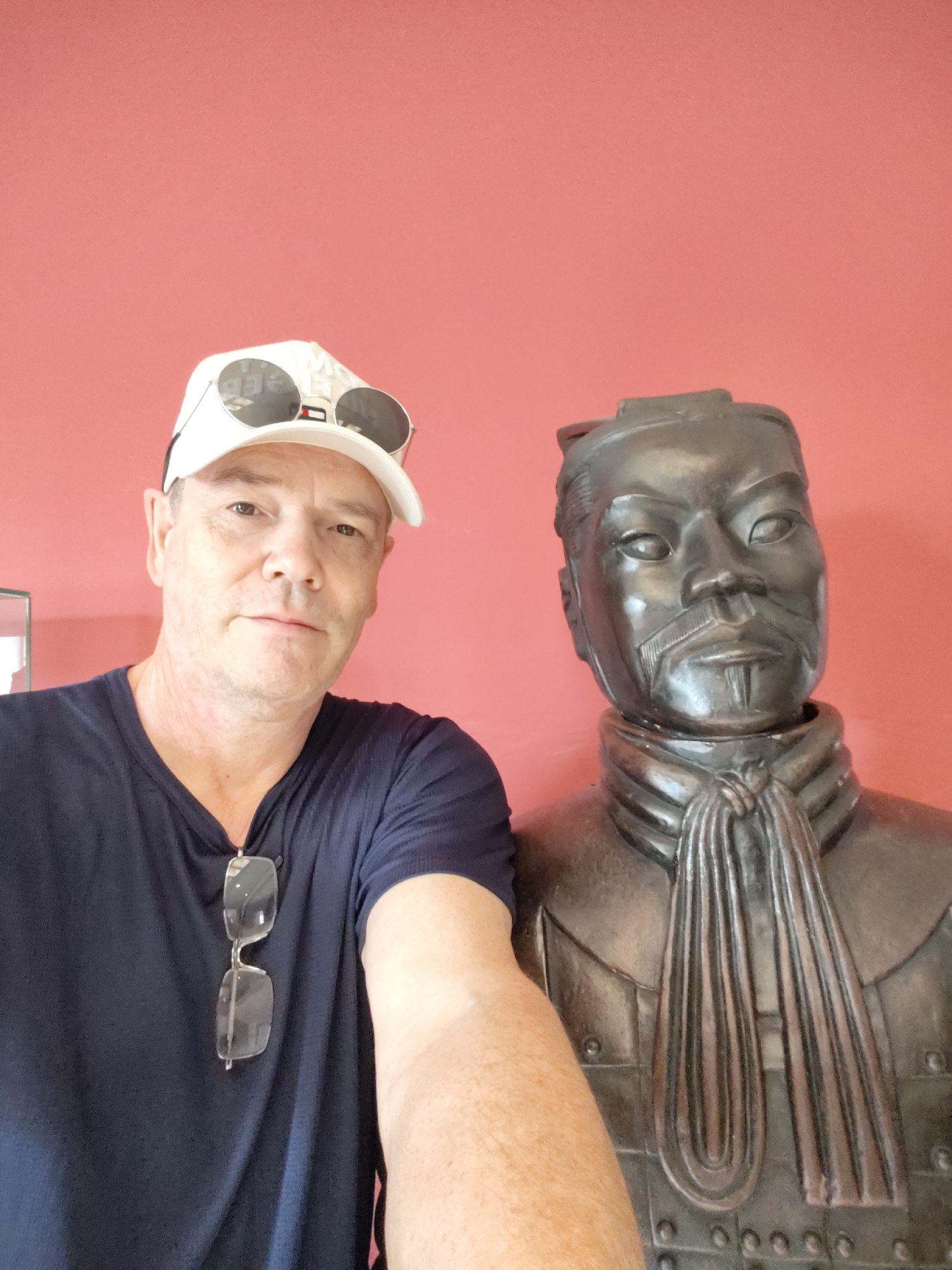 The image shows a man and a statue in front of a red wall. The man is wearing a navy blue t-shirt, a white baseball cap with sunglasses on top, and has glasses hanging from his shirt. He is looking at the camera, and his right arm is extended. To his right is a dark, bronze-colored statue of a Terracotta Warrior, depicted from the chest up, wearing armor and a distinctive scarf. The statue's face has strong features, including a mustache and a goatee. The lighting suggests an indoor setting, possibly a museum or exhibition space. The man appears to be taking a selfie with the statue. The background is a plain red wall, characteristic of many display environments in museums and cultural attractions. The location of 幸福乡, China is not specified in the image or its crops.