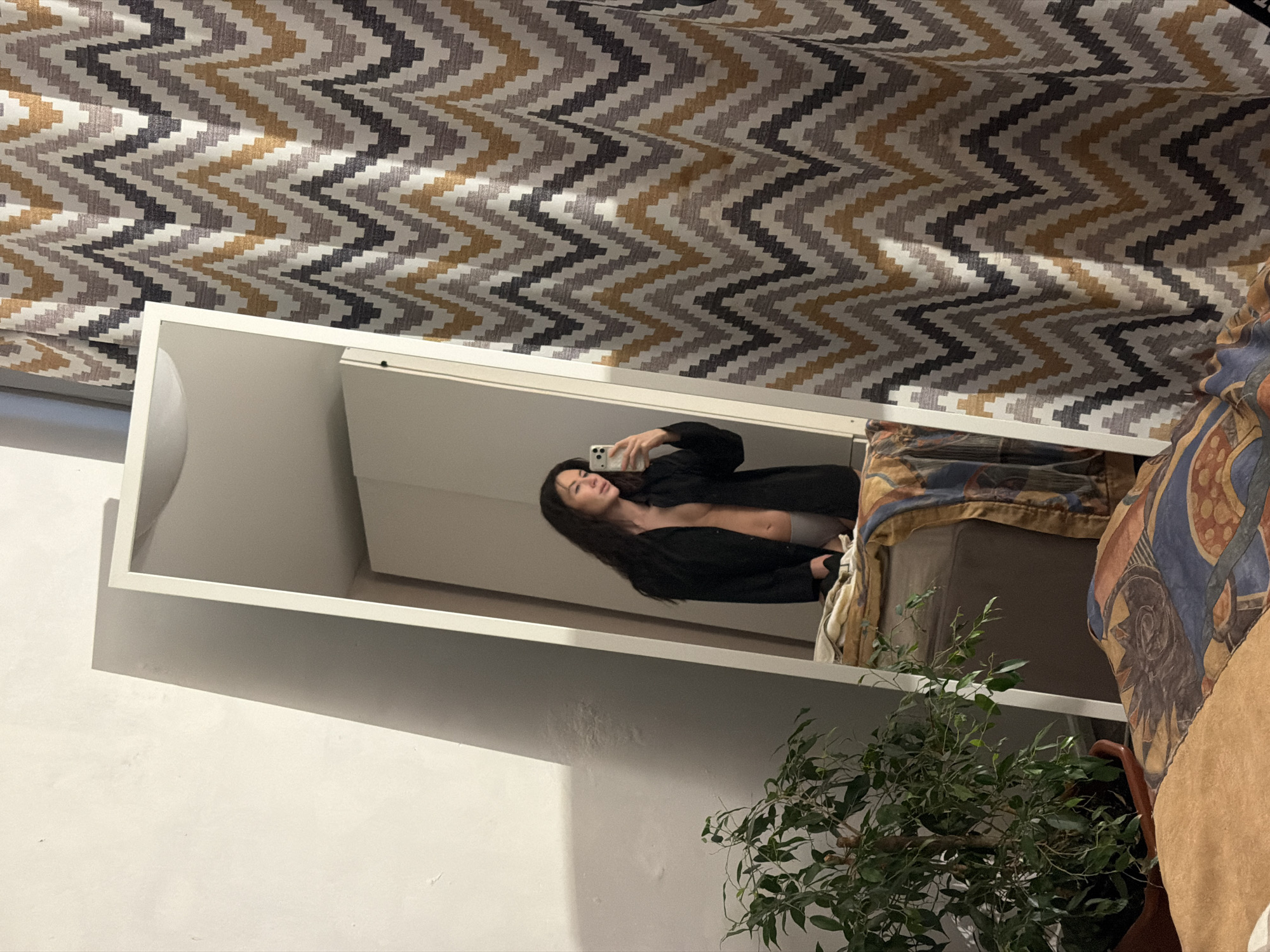 A full-length, white-framed mirror is angled within an indoor setting, reflecting a woman engaged in taking a selfie with a white smartphone. The woman has long, dark hair and is wearing an unbuttoned dark-colored shirt or blazer, revealing her bare chest and light-colored underwear bottoms. Her right hand holds the phone, angled towards the mirror.
The mirror stands between a plain, light-colored wall on the left, which exhibits a distinct diagonal shadow extending from the upper left, and a chevron-patterned curtain on the right. This curtain features a repeating zigzag design in shades of off-white, light grey, dark grey, and a muted gold/ochre.
In the lower left foreground, a potted green plant with numerous small leaves is visible in a brown pot. Below the mirror, a neutral-toned (light brown/tan) blanket covers a surface, likely a bed or sofa, with a more elaborately patterned blanket, featuring abstract designs in blue, orange, and brown, draped over it. A portion of a white ceiling with a recessed light fixture is visible in the upper reflection above the woman. No specific visual identifiers within the image directly indicate its precise location in Perm, Russia. The scene depicts a private, lived-in interior space.