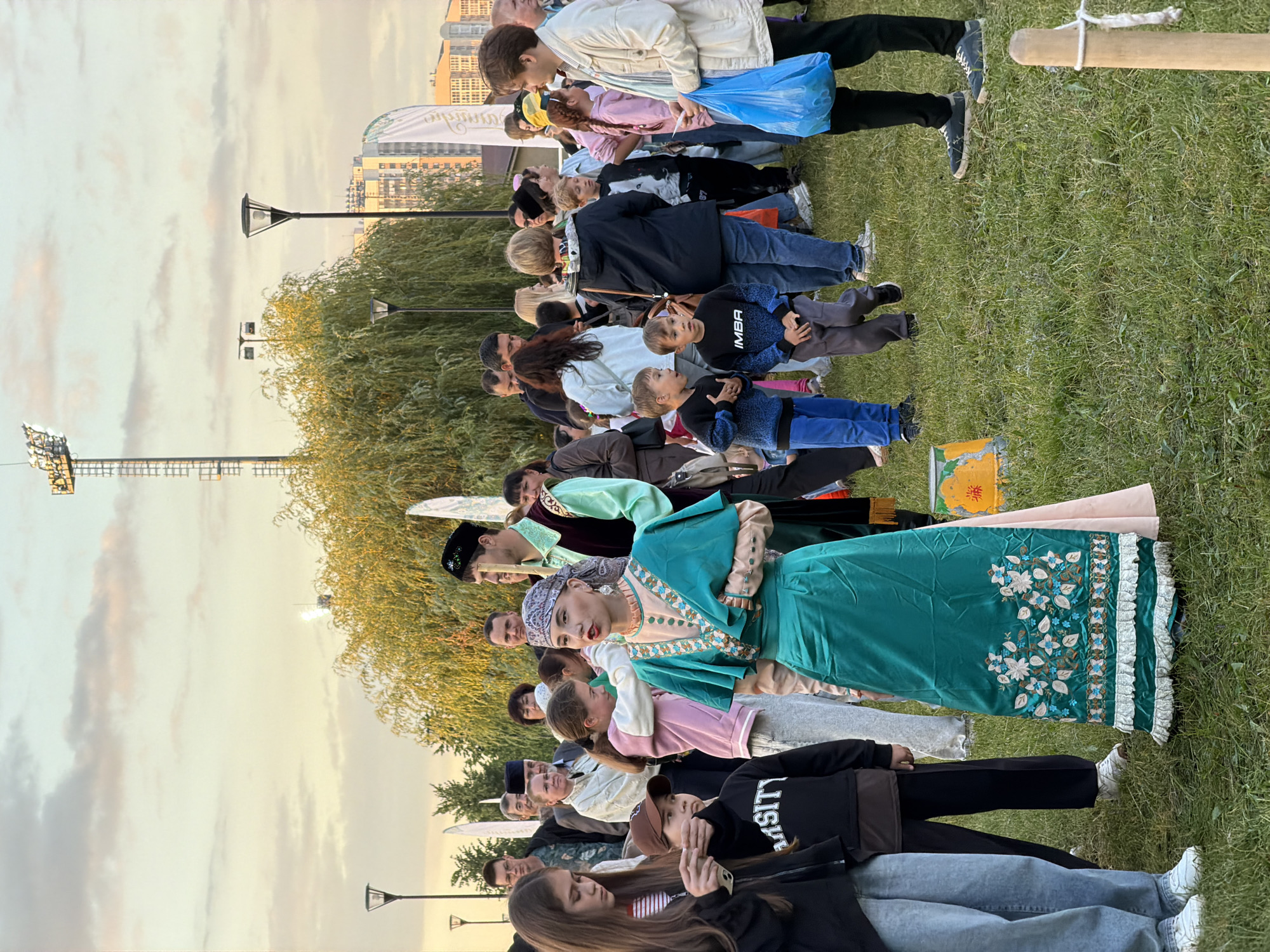 An outdoor gathering is depicted in an urban park. The sky is a pale blue with scattered clouds, and the sun appears to be low in the sky, casting a warm glow. A large, luminous floodlight tower stands in the background, along with several modern buildings. The foreground is dominated by a grassy field where a diverse crowd of people, including adults and children, are milling about. A woman in traditional attire, a teal dress adorned with intricate embroidery and a headscarf, is prominently featured in the center. She is surrounded by other individuals, some of whom are looking at her, while others are engaged in conversations or observing their surroundings. A young girl in a pink hoodie stands next to the woman, and to the left, a girl with long brown hair holds a smartphone. In the background, men and women are seen in casual clothing. The overall atmosphere suggests a community event or festival.