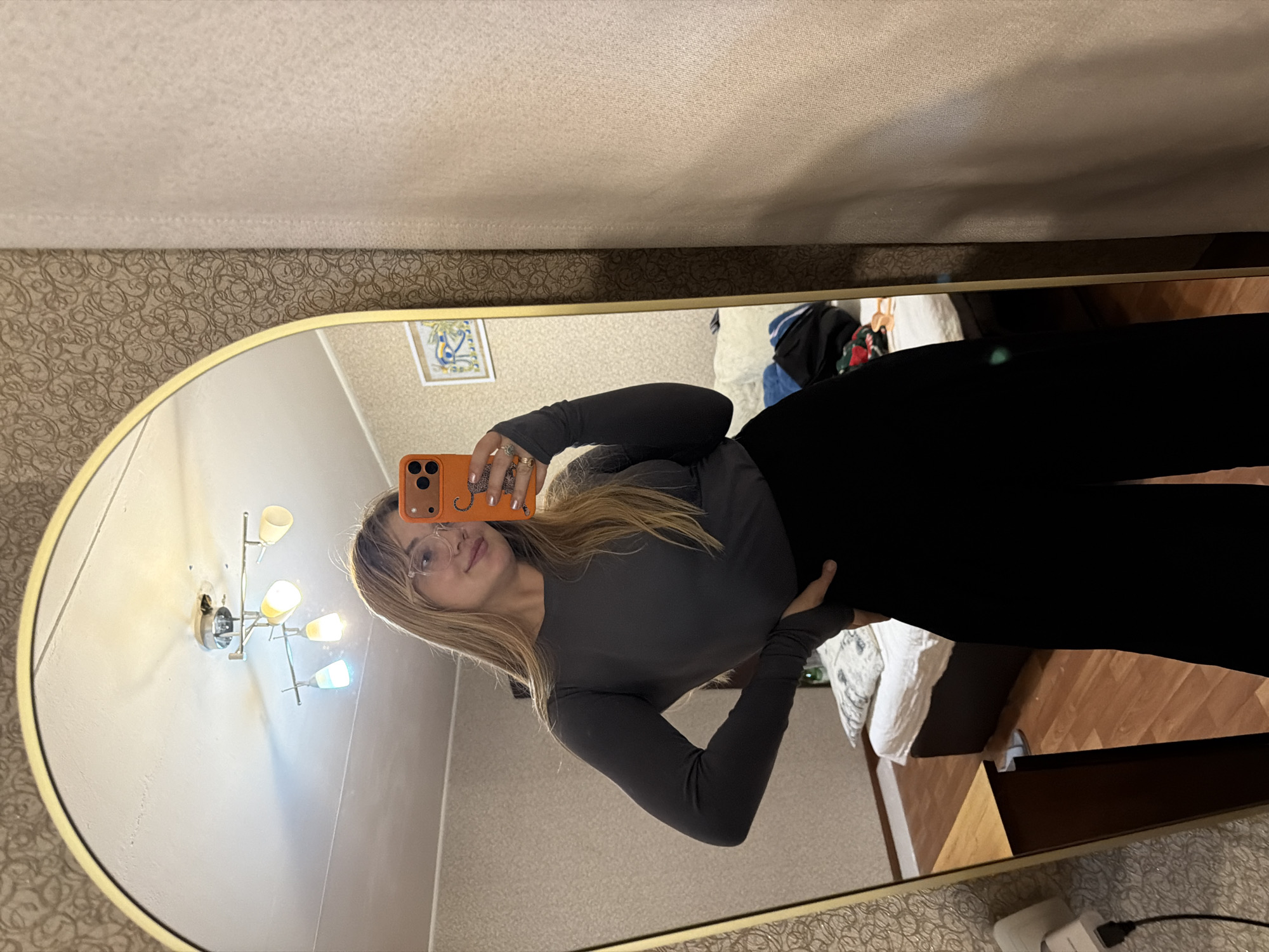A young woman is taking a selfie in a mirror. The mirror is oval-shaped with a gold frame and is reflecting a room. The woman has long blonde hair and is wearing glasses, a gray long-sleeved shirt, and black pants. She is holding an orange phone with a leopard print case. The background of the reflection shows a room with light-colored walls, a patterned wallpaper border, and a chandelier. There is also a bed with white sheets and a wooden floor visible in the reflection. The location is likely in Naberezhnye Chelny, Russia, based on the user's request.